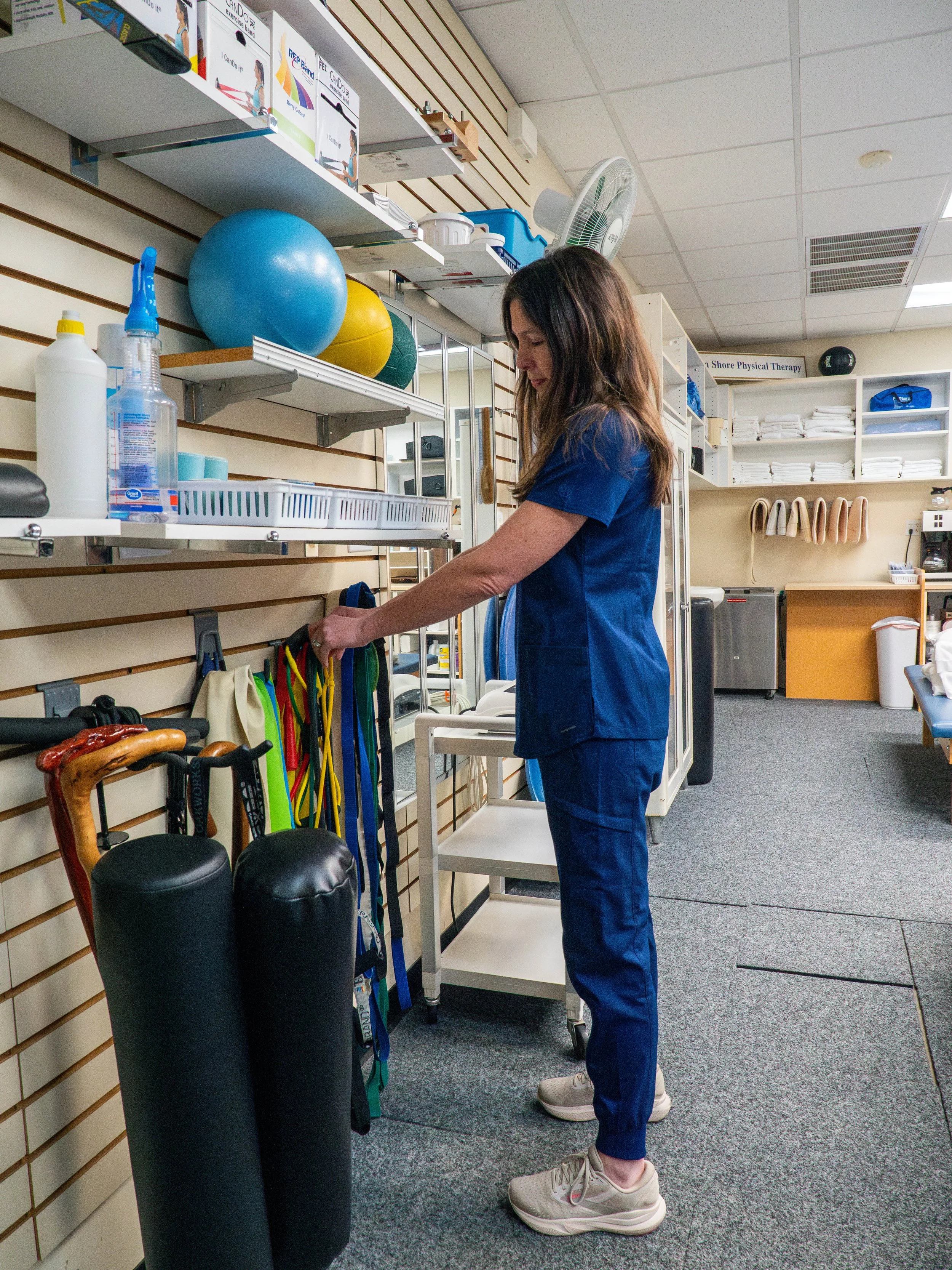A woman in blue medical scrubs organizing resistance bands on a wall in a physical therapy or medical supply room.