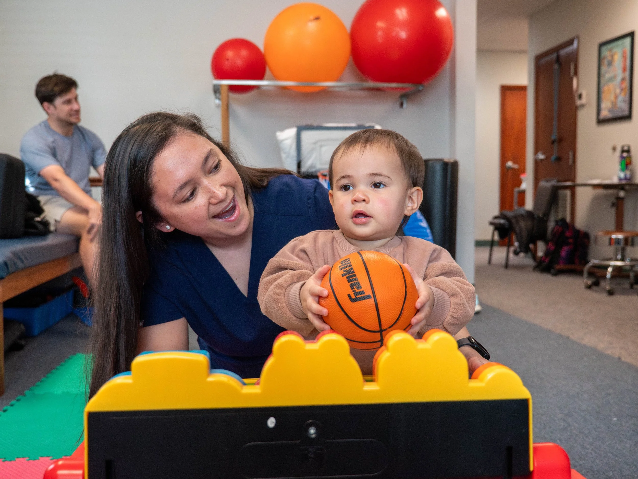 A young boy holding an orange basketball while leaning on a yellow and black toy basketball set, with a woman kneeling beside him, in a therapy or activity room with colorful therapy balls and equipment in the background.