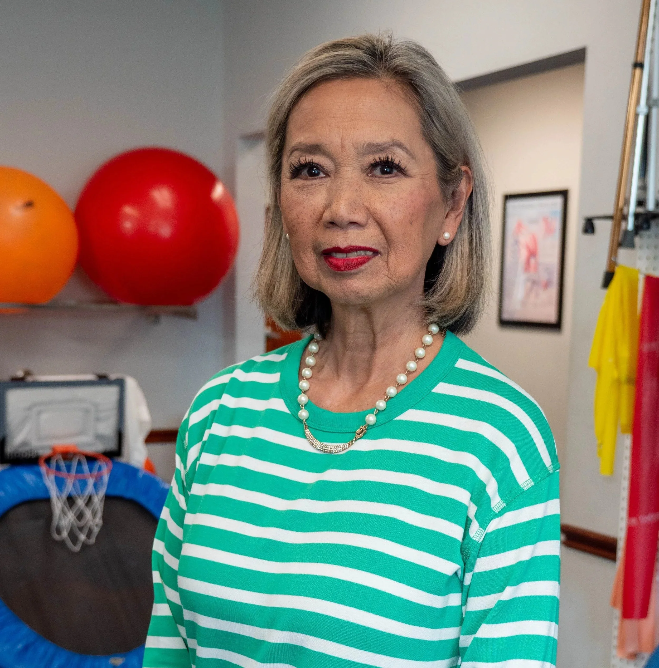 An older woman with gray hair, wearing a green and white striped shirt, red lipstick, pearl necklace, and pearl earrings, standing indoors near sports equipment and exercise balls.