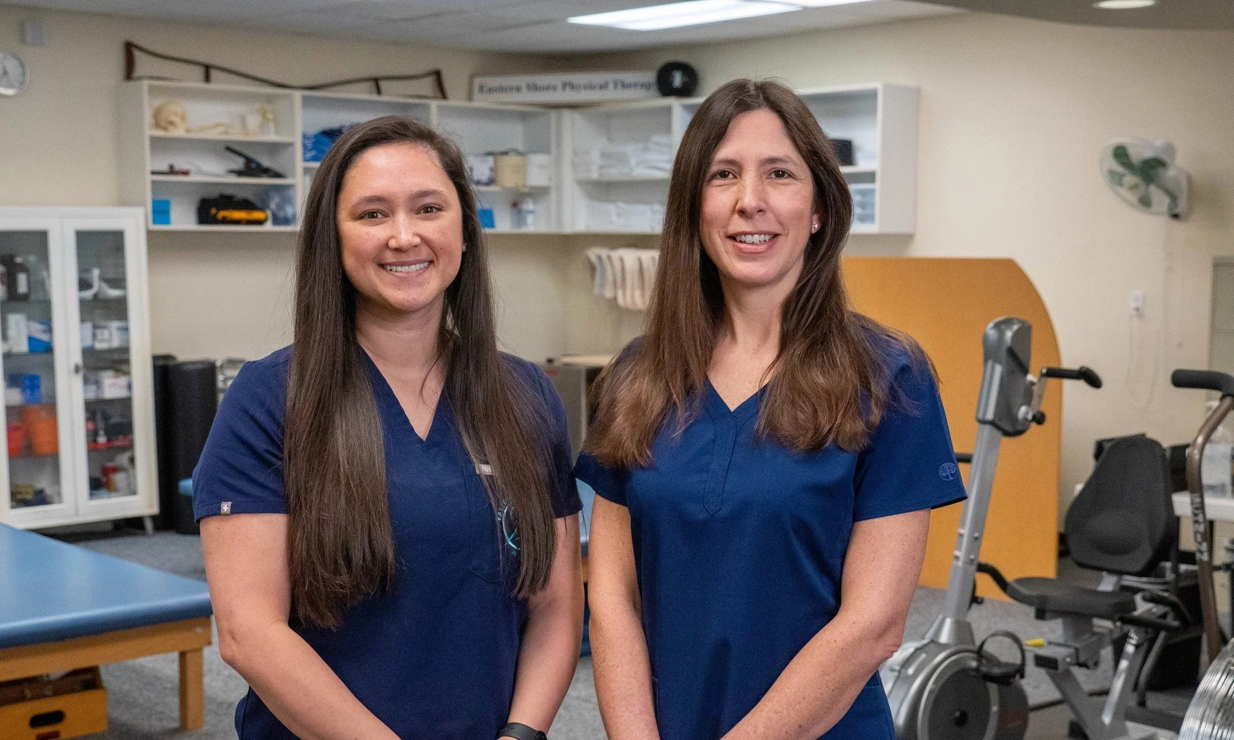 Two women wearing blue medical scrubs standing inside a medical office or therapy room, with exercise equipment and shelves in the background.