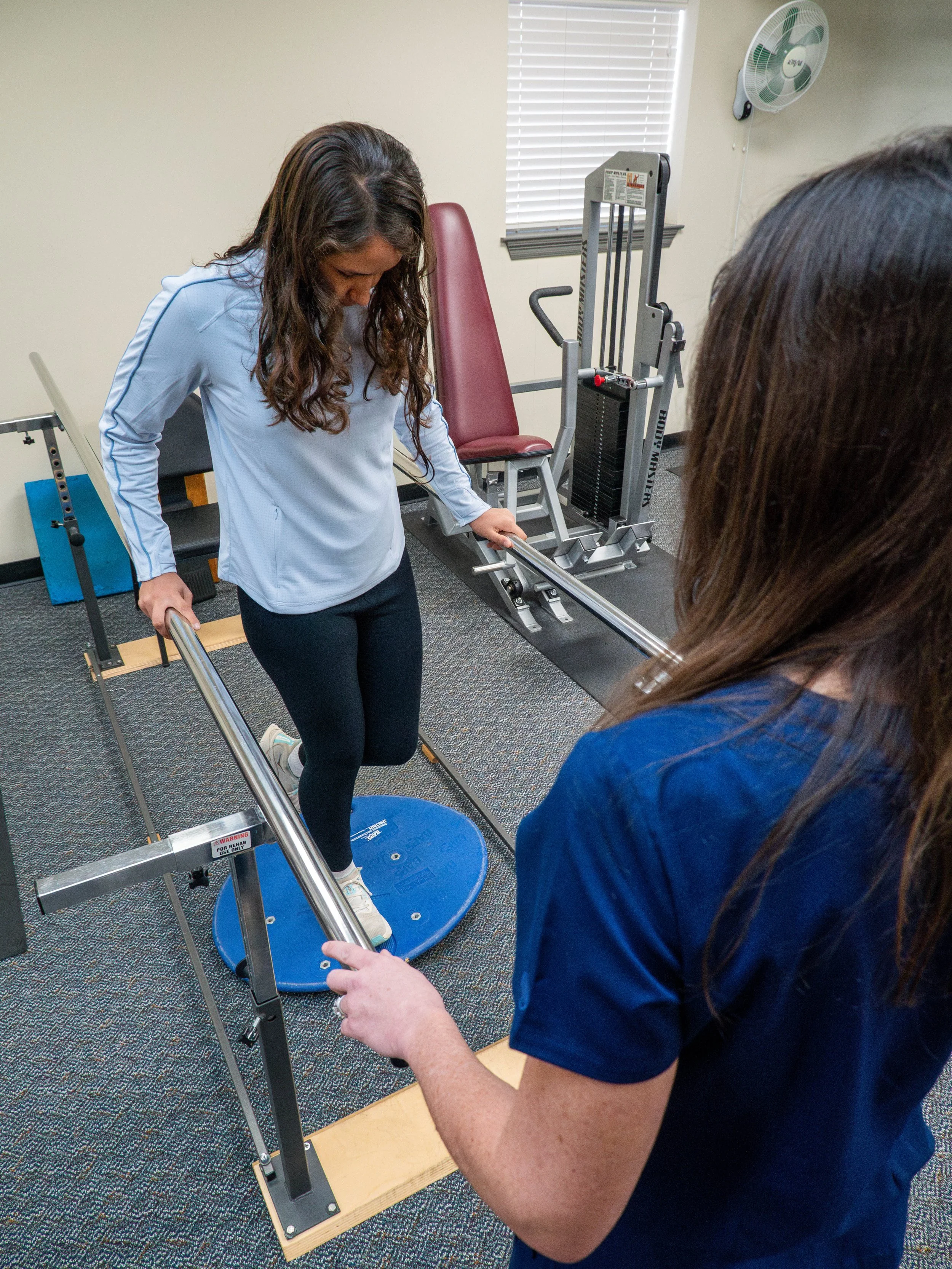 Physical therapist assisting a woman in balancing on a balance disc while holding onto parallel bars in a rehabilitation setting.