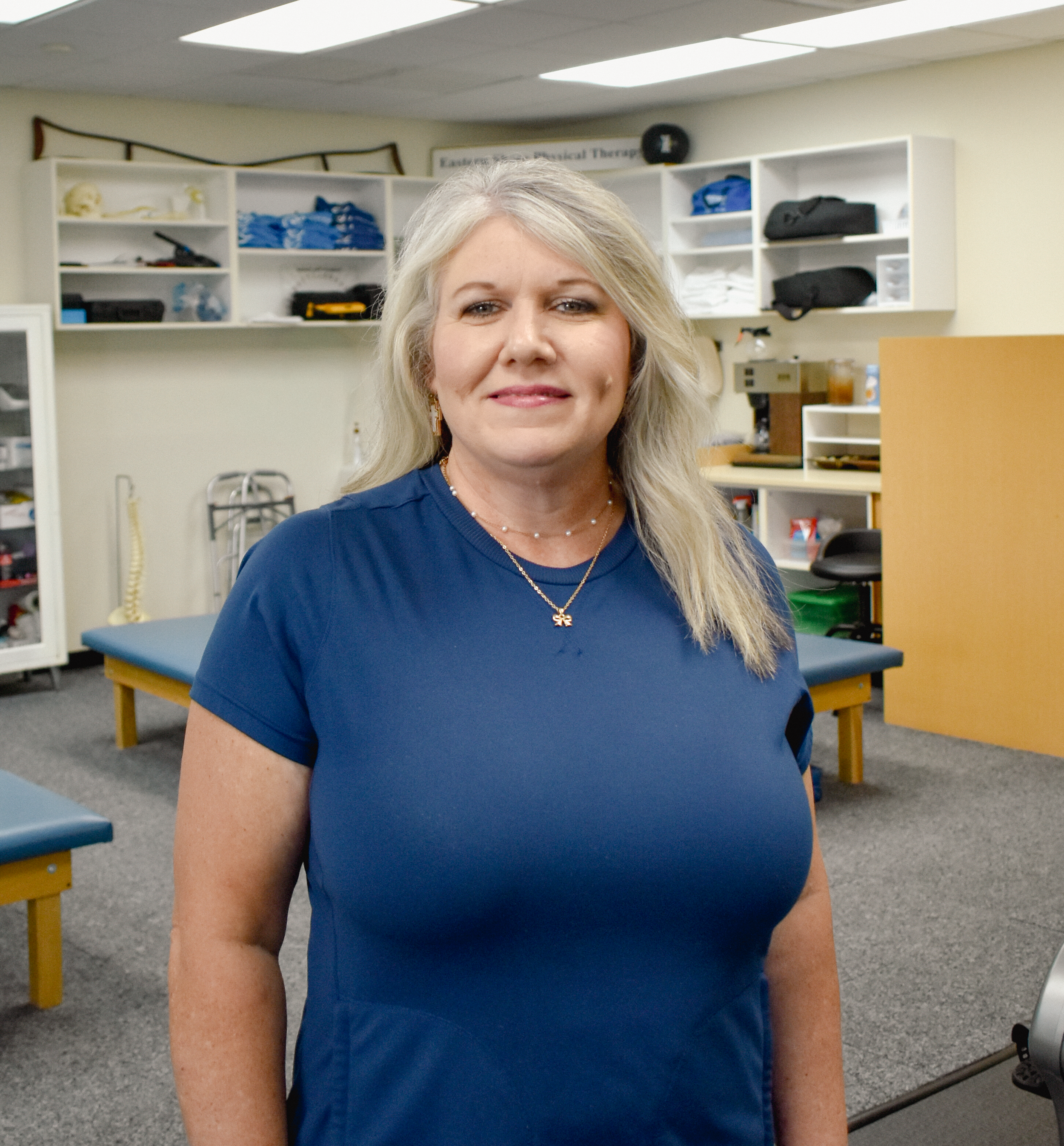 A woman with long blonde hair wearing a blue top and jewelry, standing in a medical or therapy room with shelves, medical equipment, and tables in the background.