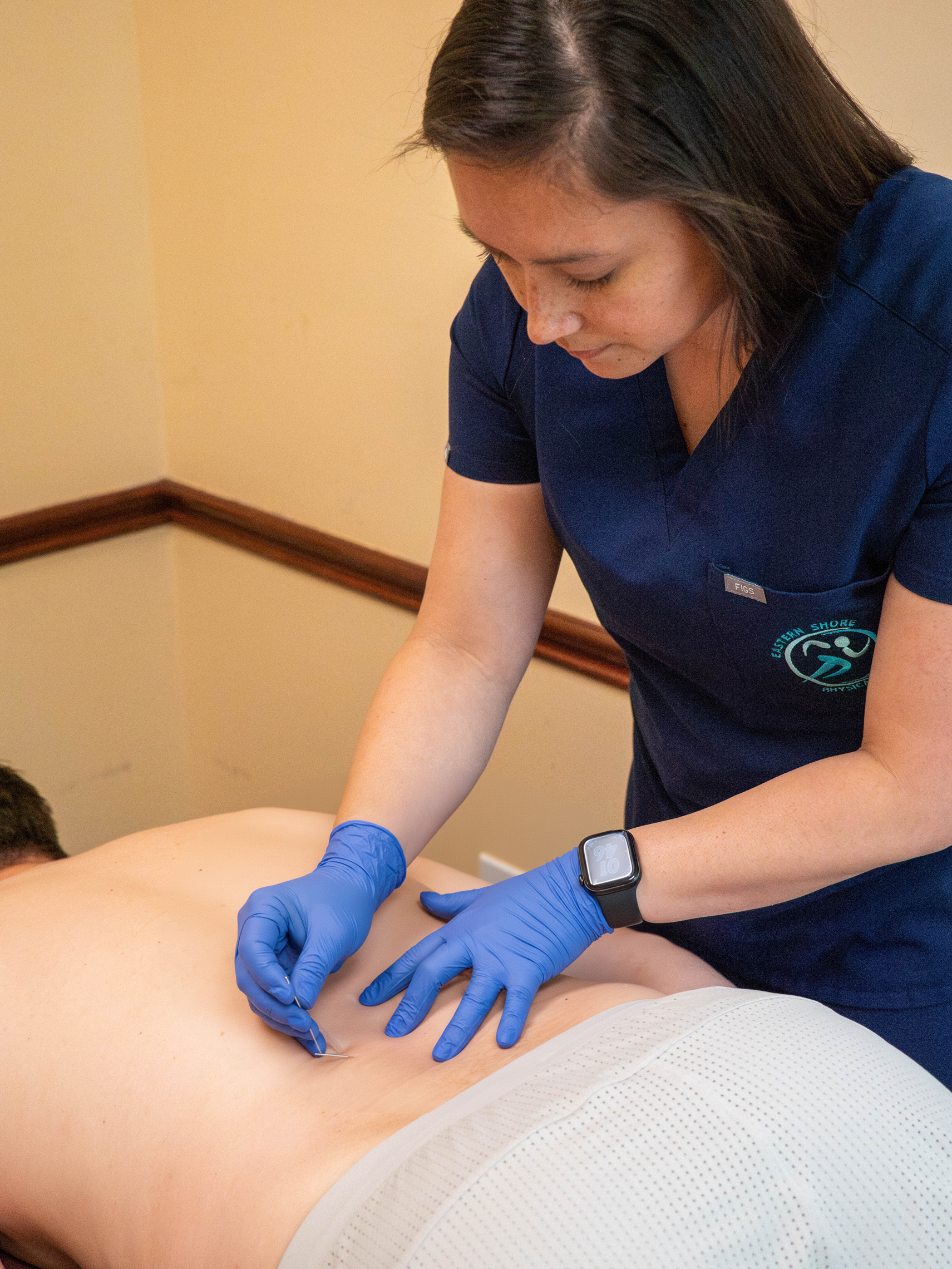 A healthcare worker in blue scrubs and gloves performs acupuncture on a patient lying face down on a treatment table.