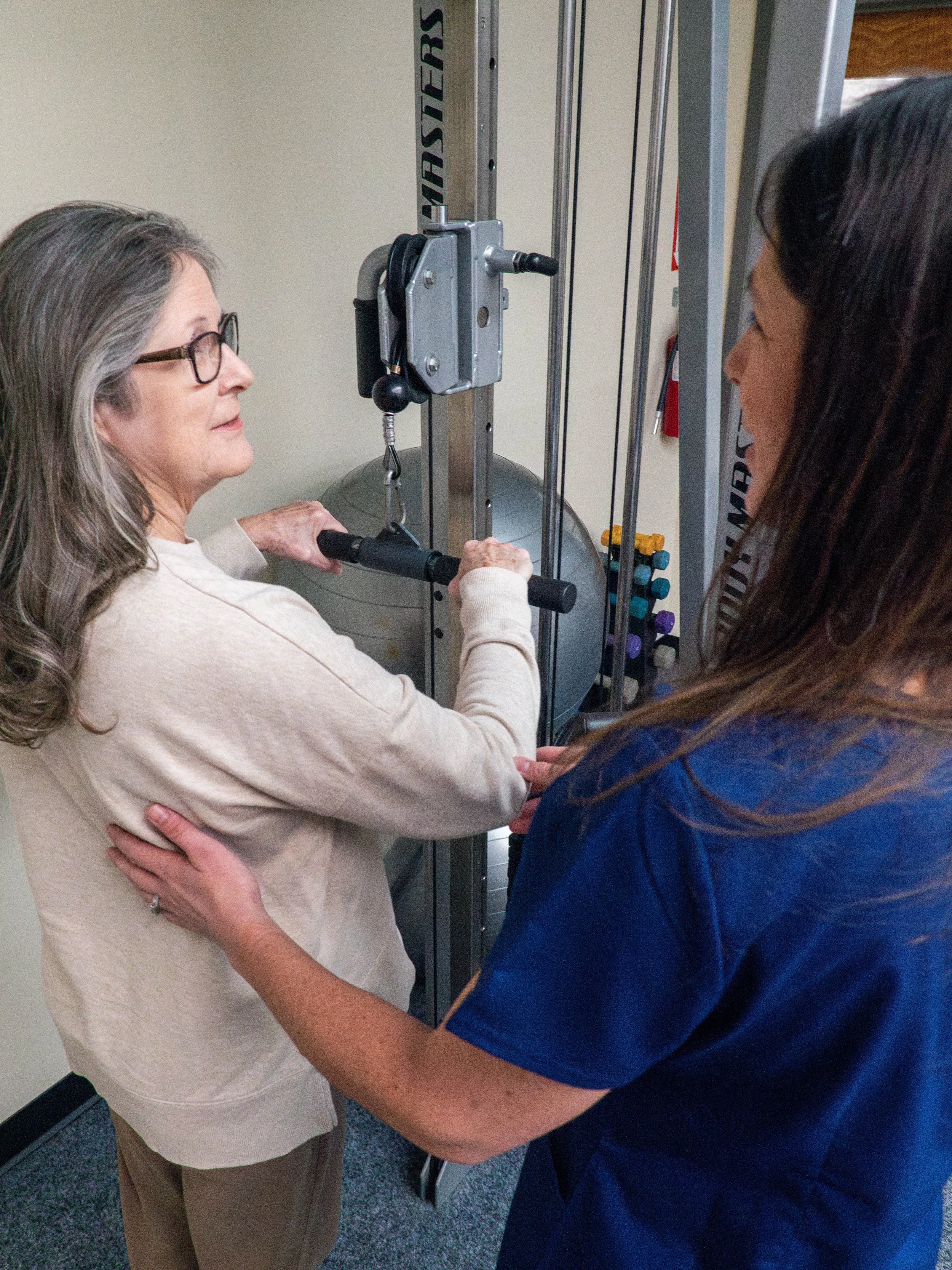 A senior woman with glasses and gray hair exercising her arm on a pulley machine in a medical or physical therapy setting, assisted by a healthcare professional in navy scrubs.