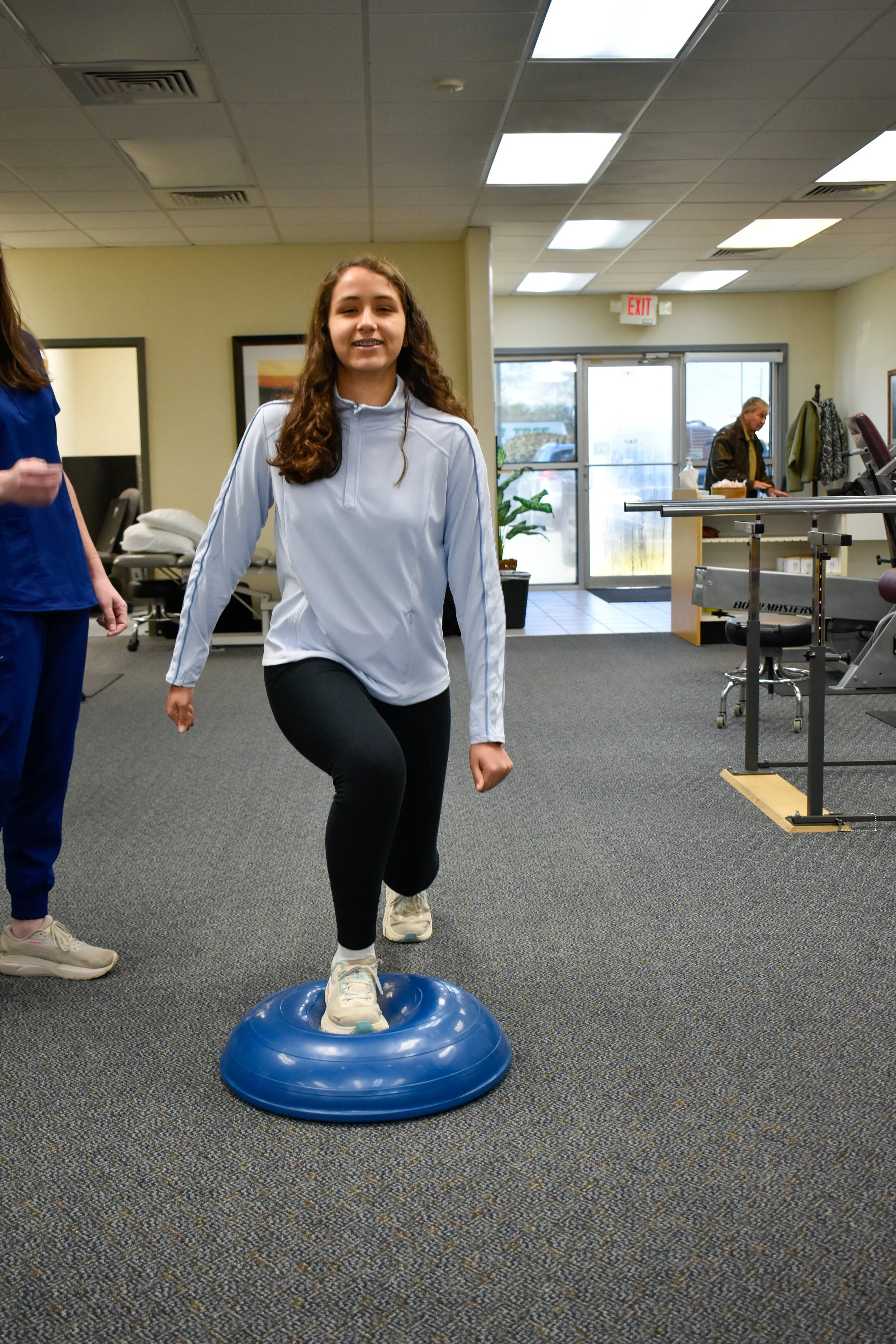 Young woman balancing on a blue exercise disc in a physical therapy or gym setting, with a trainer nearby.