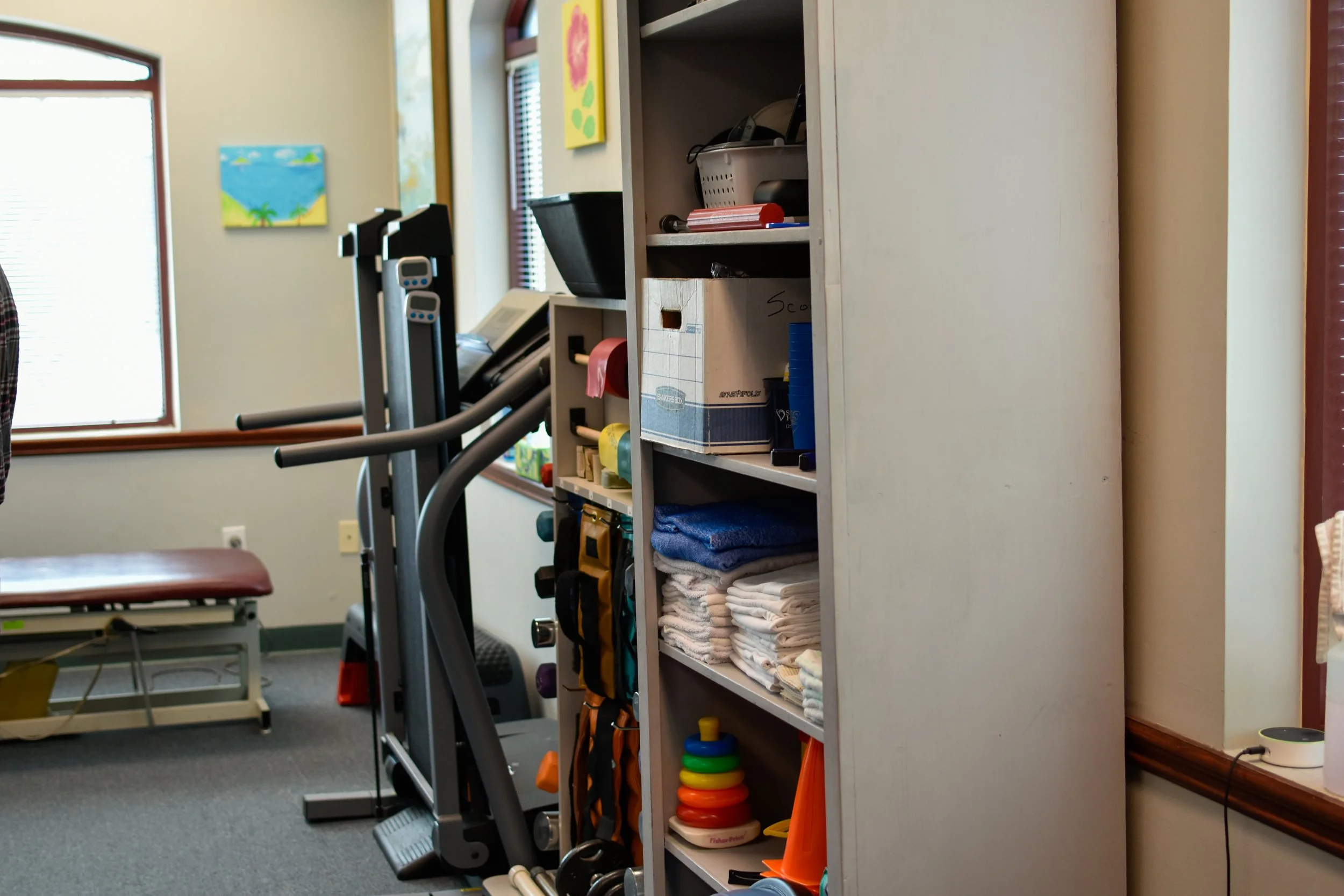 A shelf filled with rolled and folded towels, exercise equipment, and colorful toys in a medical or therapy room with a bench and exercise machines nearby.