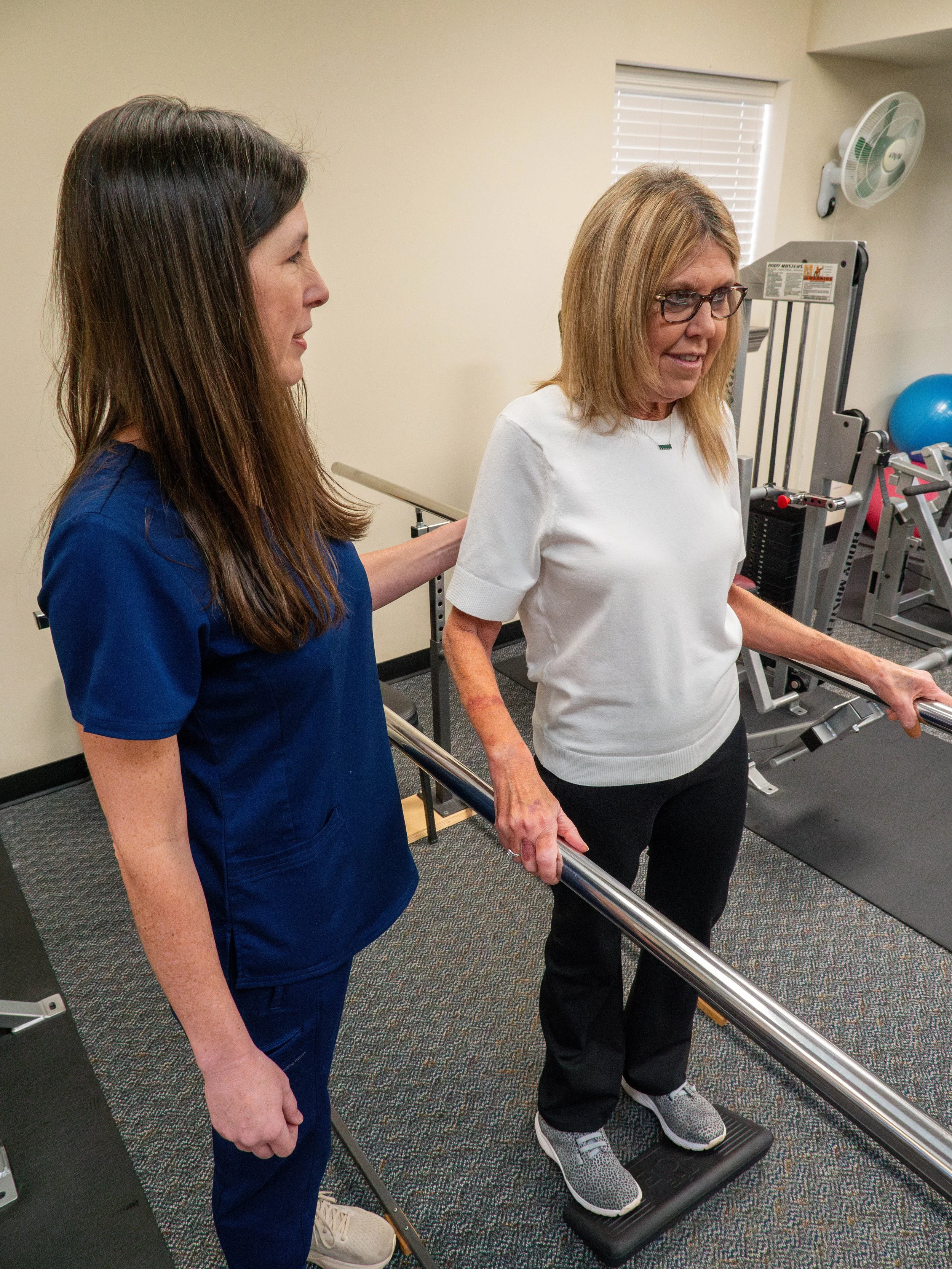 A woman in a white t-shirt and black pants standing on a treadmill, holding onto the handrails, being assisted or guided by a woman in medical scrubs in a therapy or rehabilitation setting with exercise equipment such as a blue stability ball and a standing frame in the background.
