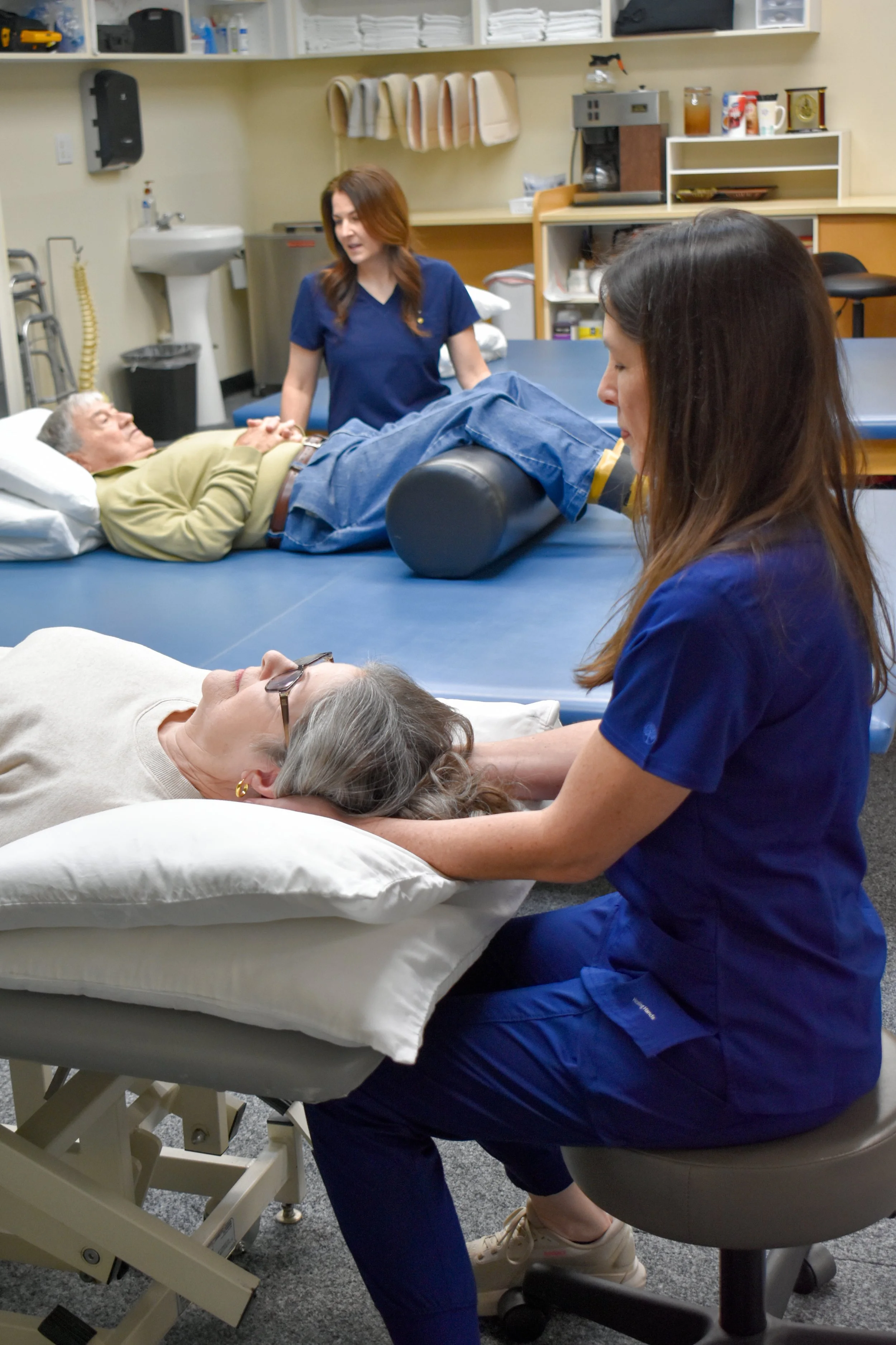 A nurse in blue scrubs is giving a woman a head massage on a bed in a healthcare setting. An elderly man is lying on a treatment table in the background.