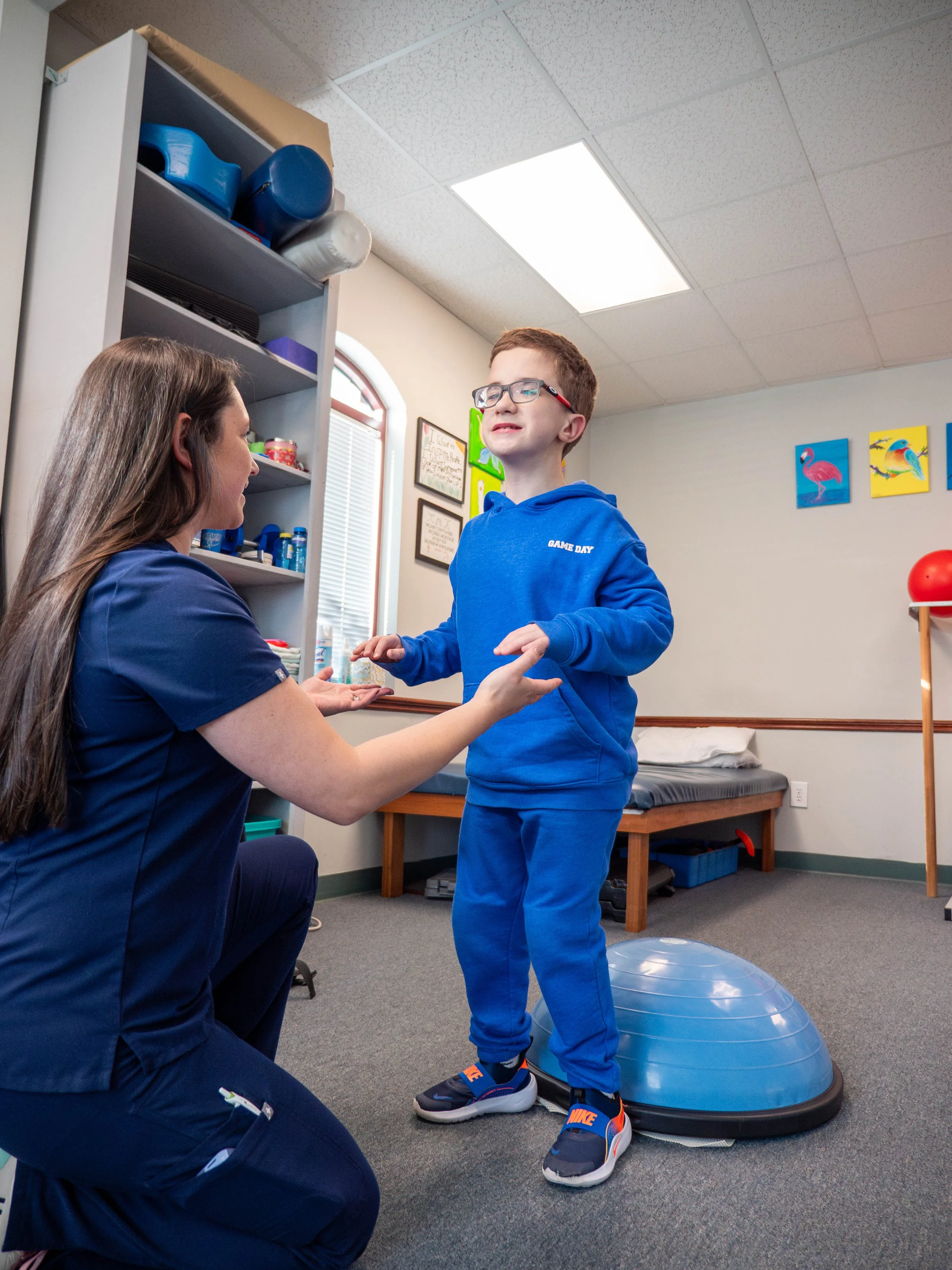 A boy in a blue hoodie and pants standing on a balance trainer while a healthcare professional in navy scrubs holds his hands, in a physical therapy or rehabilitation room.