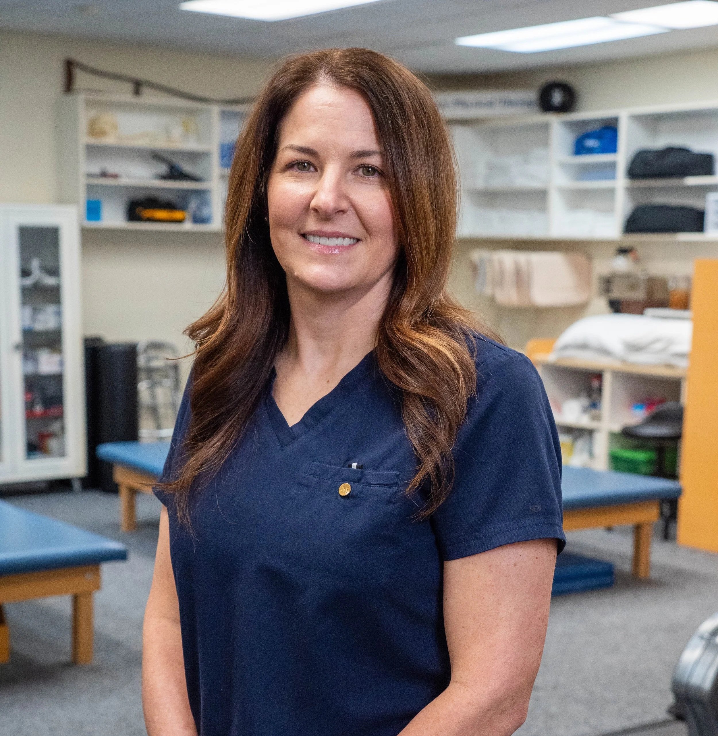 A woman wearing navy medical scrubs standing in a clinical or hospital room, smiling at the camera.