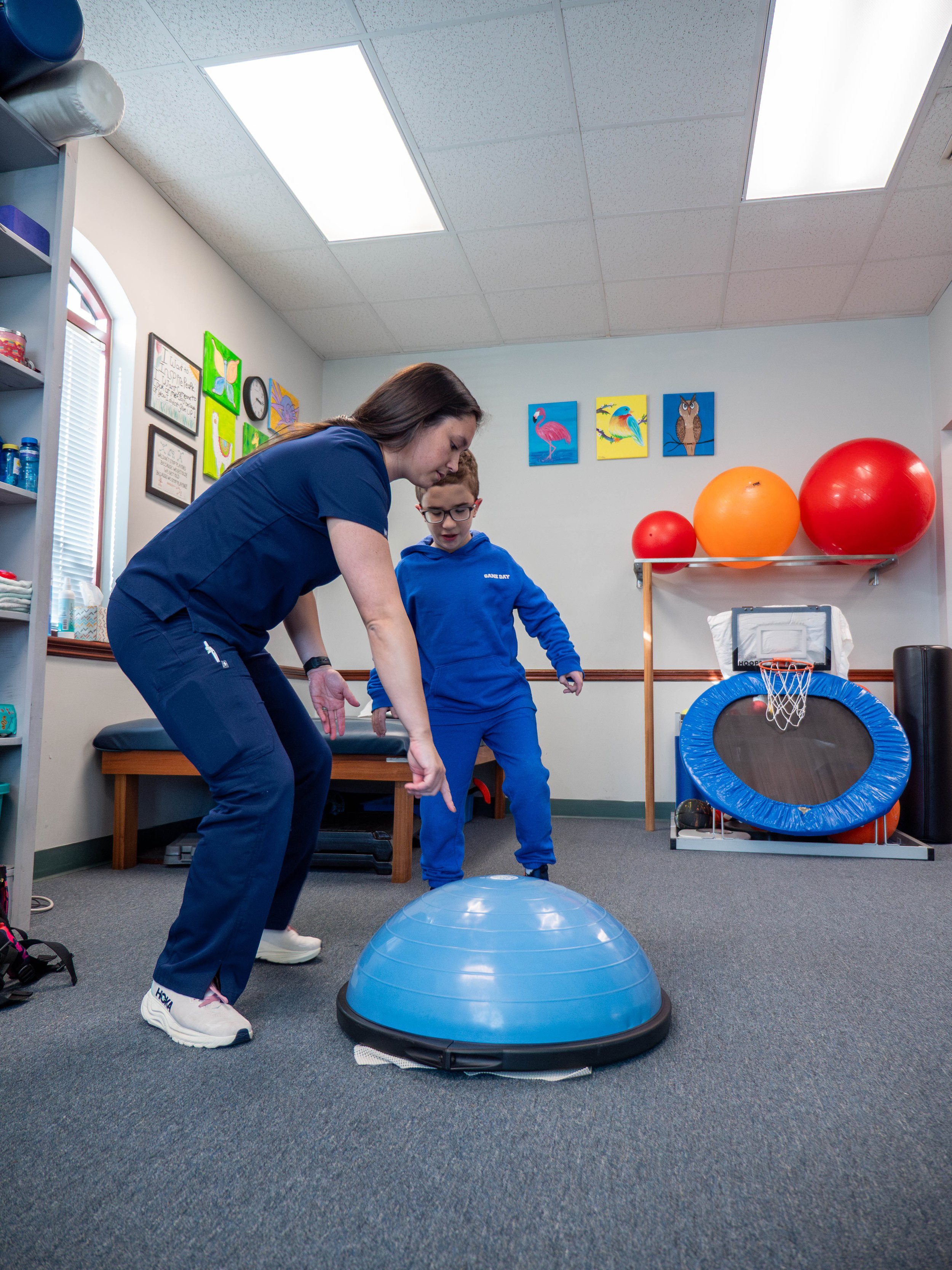 A woman and a boy in a therapy or exercise room using a balance trainer, with therapy balls and a mini basketball hoop in the background.