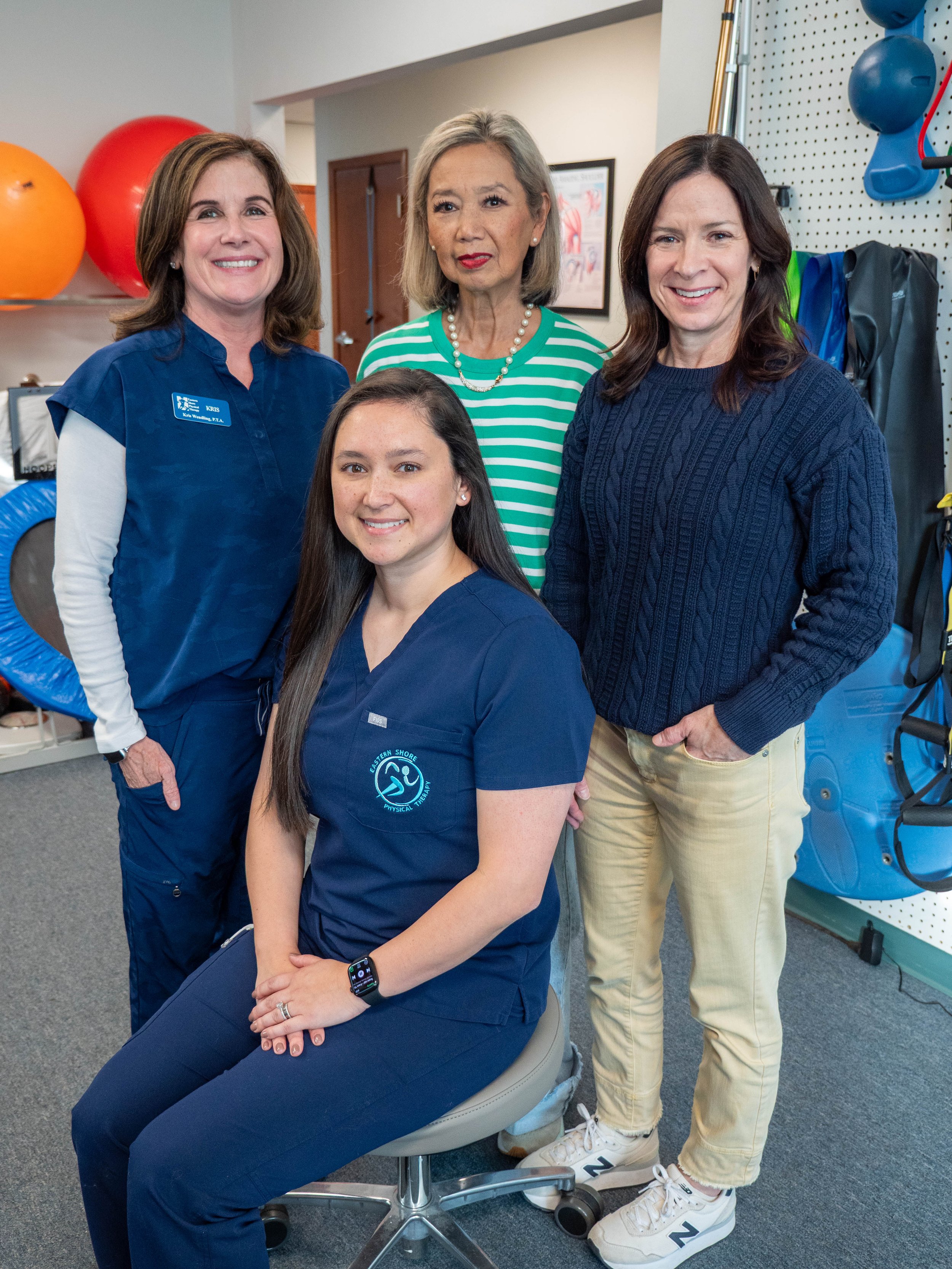 Four women, two in navy scrubs and two in casual clothing, are posing together in a medical or therapy setting with exercise equipment and a pegboard in the background.