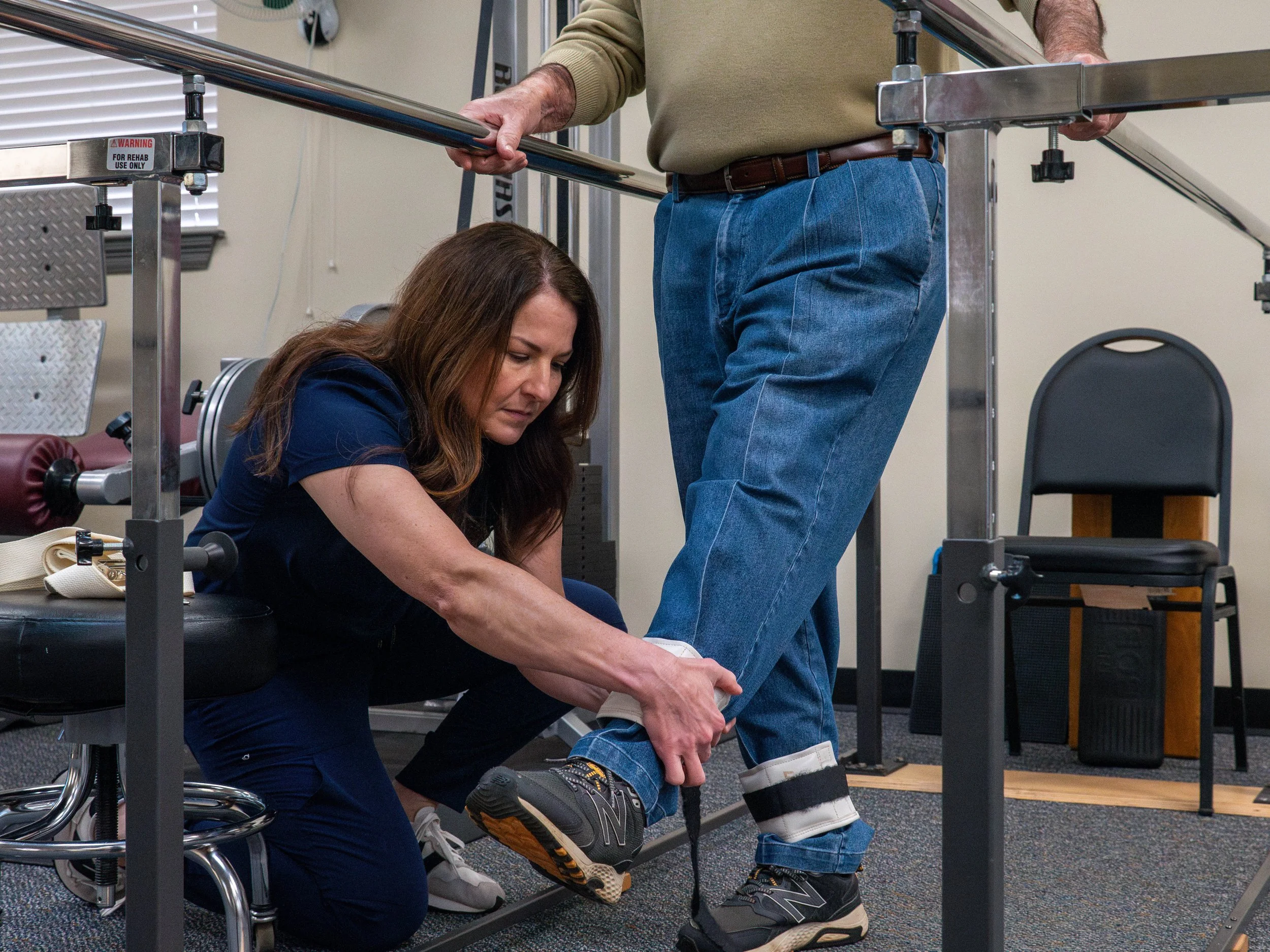A woman helping a man with his shoe in a physical therapy or rehabilitation setting, with exercise equipment and chairs in the background.