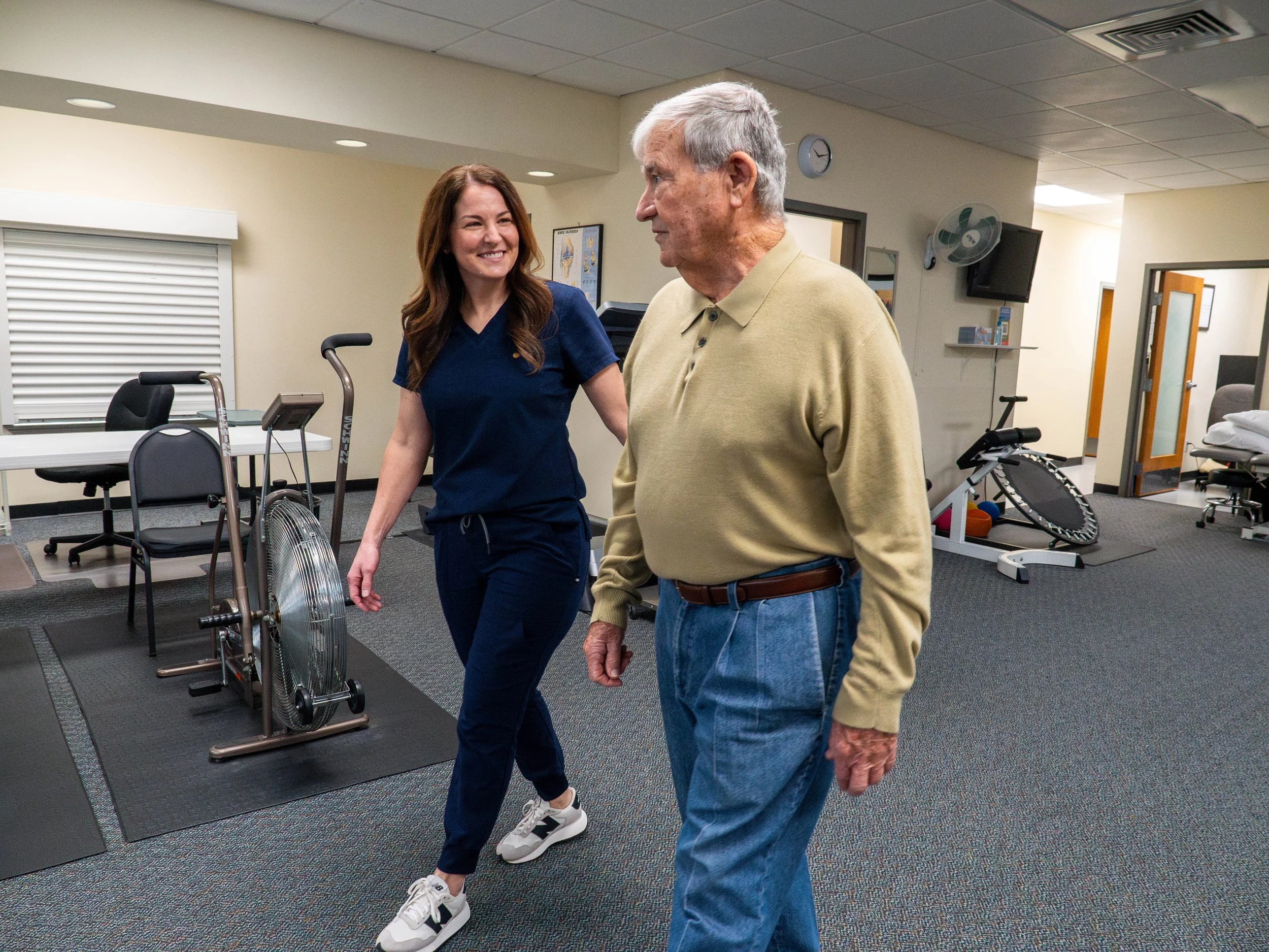 A smiling female healthcare professional walks with an elderly man in a medical or physical therapy office.