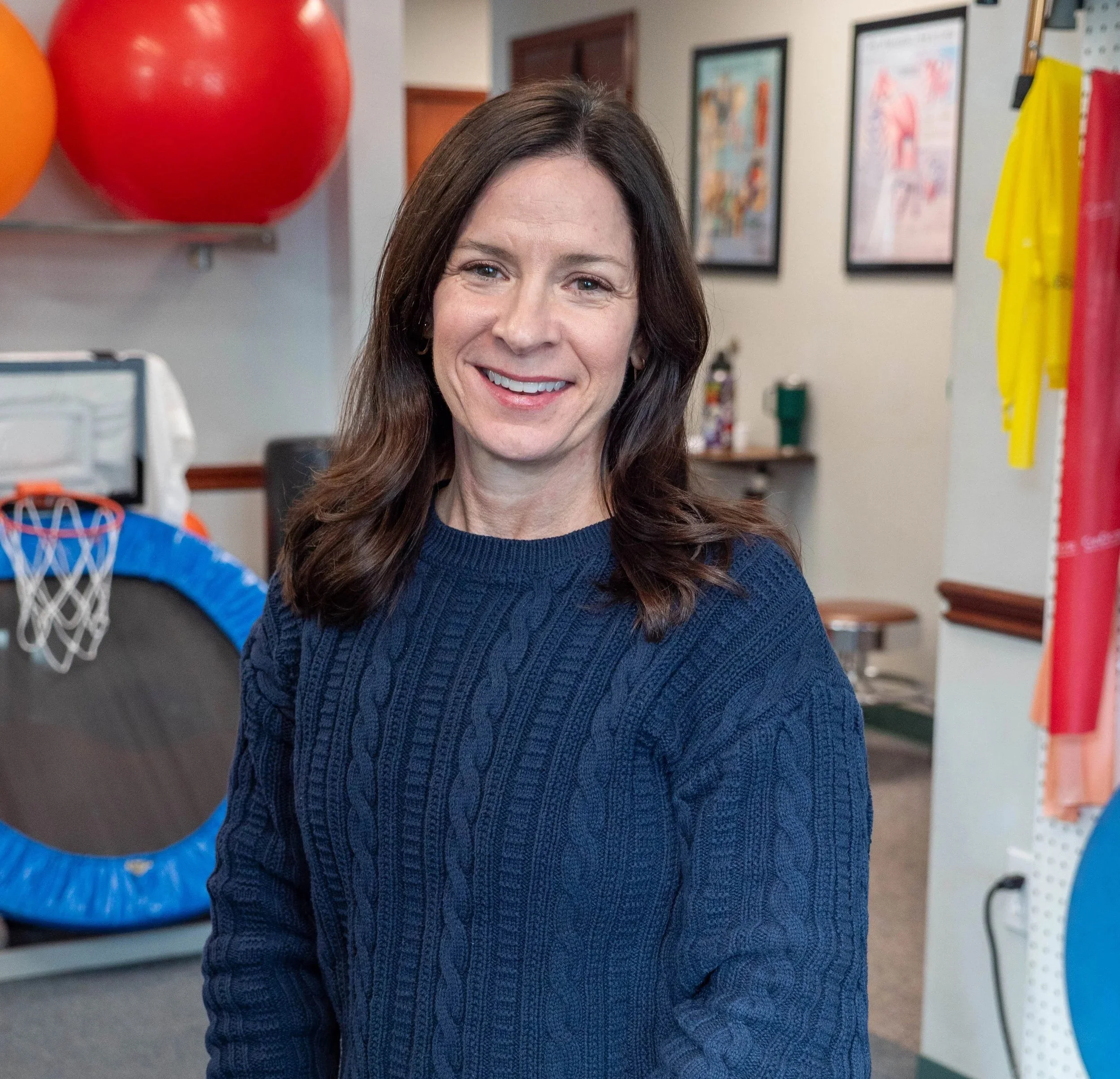 A woman with long brown hair, wearing a dark blue knitted sweater, smiling in an office or classroom environment. In the background, there are framed posters on the wall, and a basketball hoop with a blue net and orange backboard is visible on the left side of the image.