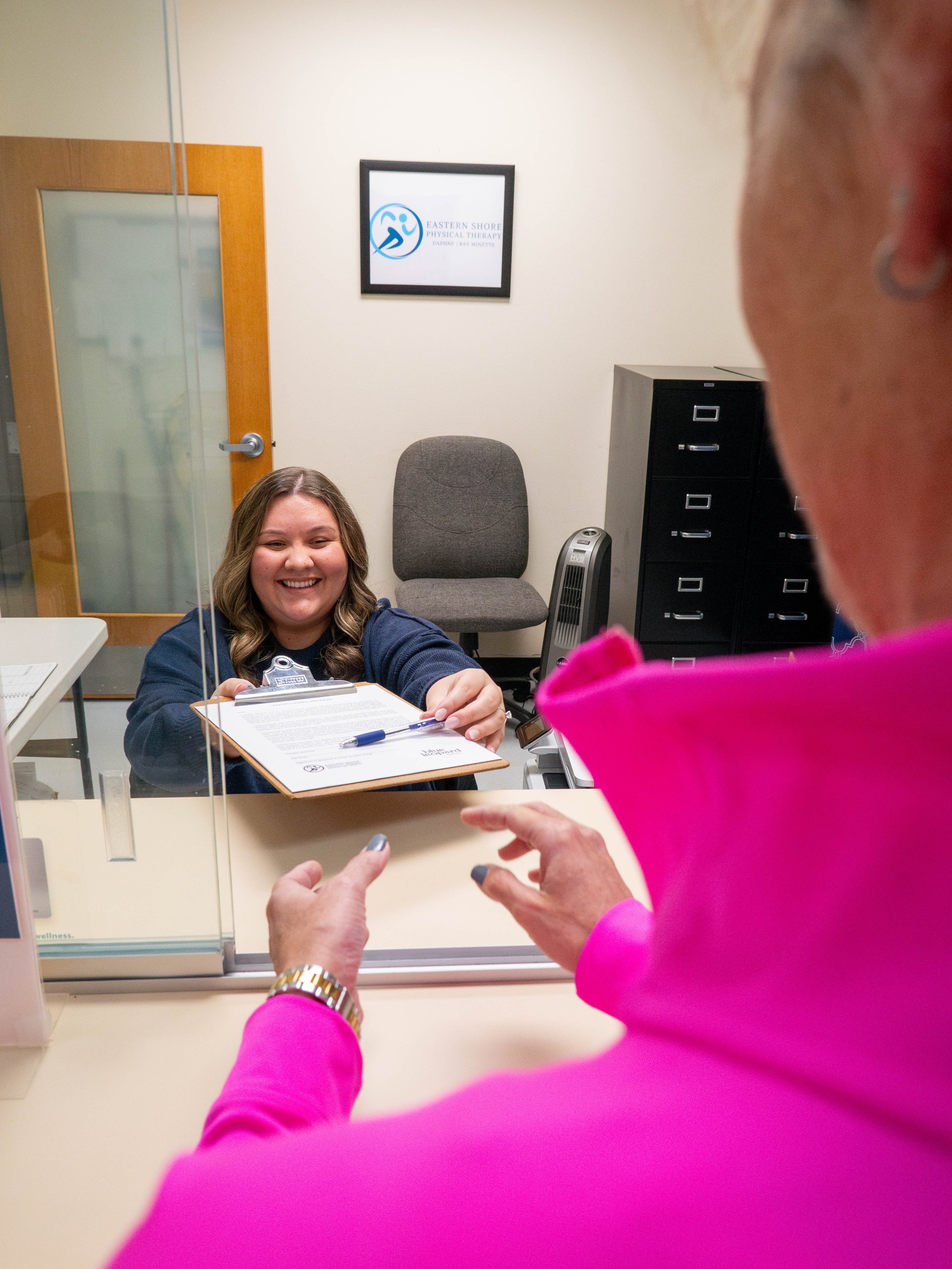 A woman smiling and handing a clipboard to a person behind a glass window in an office setting.
