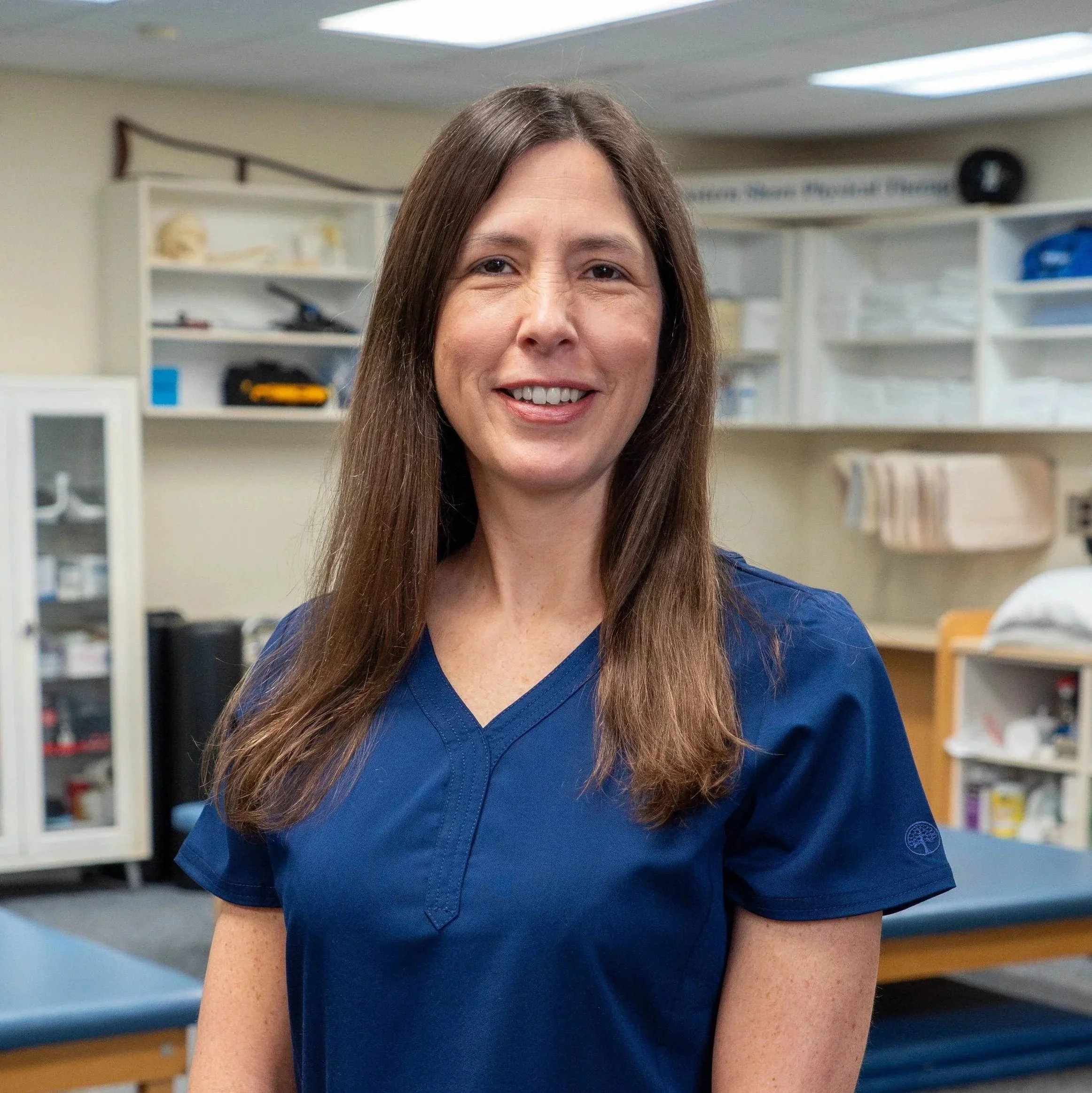 A woman with long brown hair wearing blue medical scrubs, standing in a medical or clinical setting with shelves and medical supplies in the background.