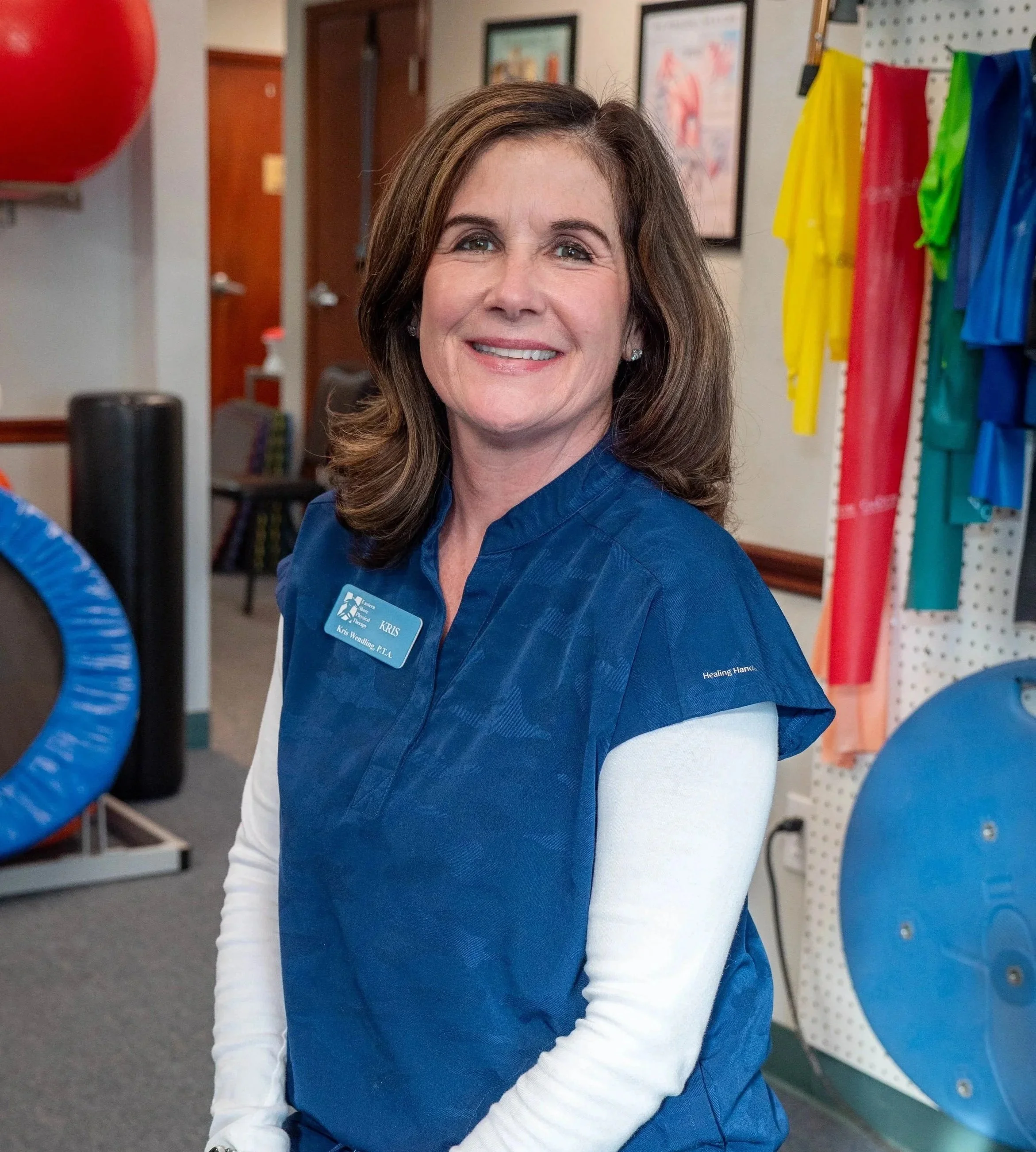 A woman smiling, wearing a blue name tag and a blue shirt, standing in a room with colorful athletic equipment and framed pictures on the wall.
