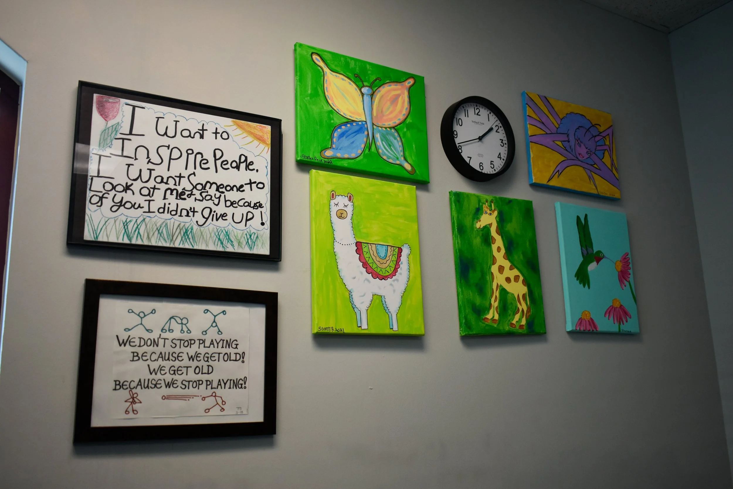 Wall decorated with colorful artwork, including paintings of a butterfly, llama, giraffe, fairy, and hummingbird, along with two motivational quotes and a round clock.