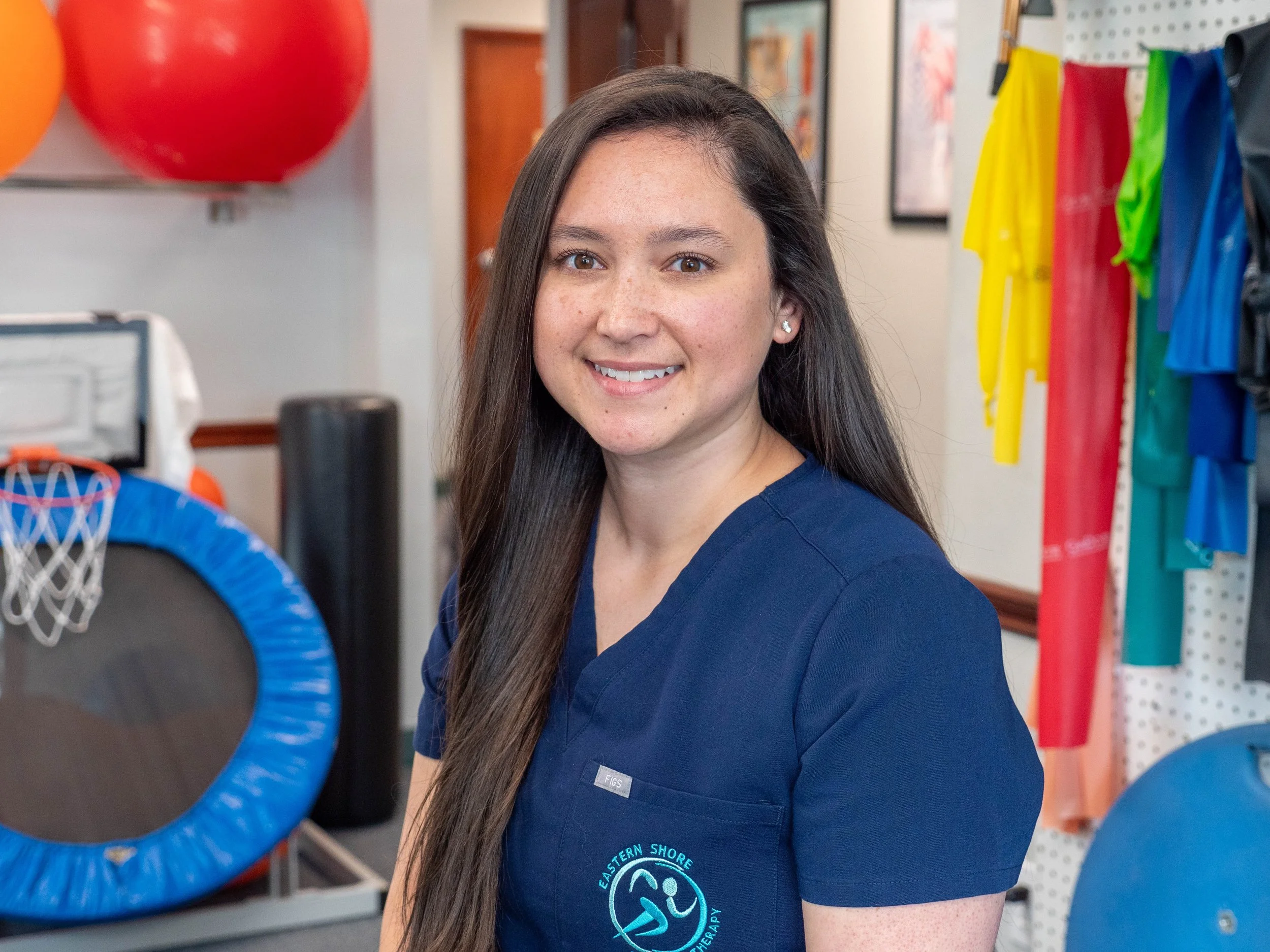 A young woman with long dark hair and a blue medical uniform with a logo standing in a sports therapy or physical therapy clinic, surrounded by colorful sports equipment including small basketball hoops and hanging sportswear.