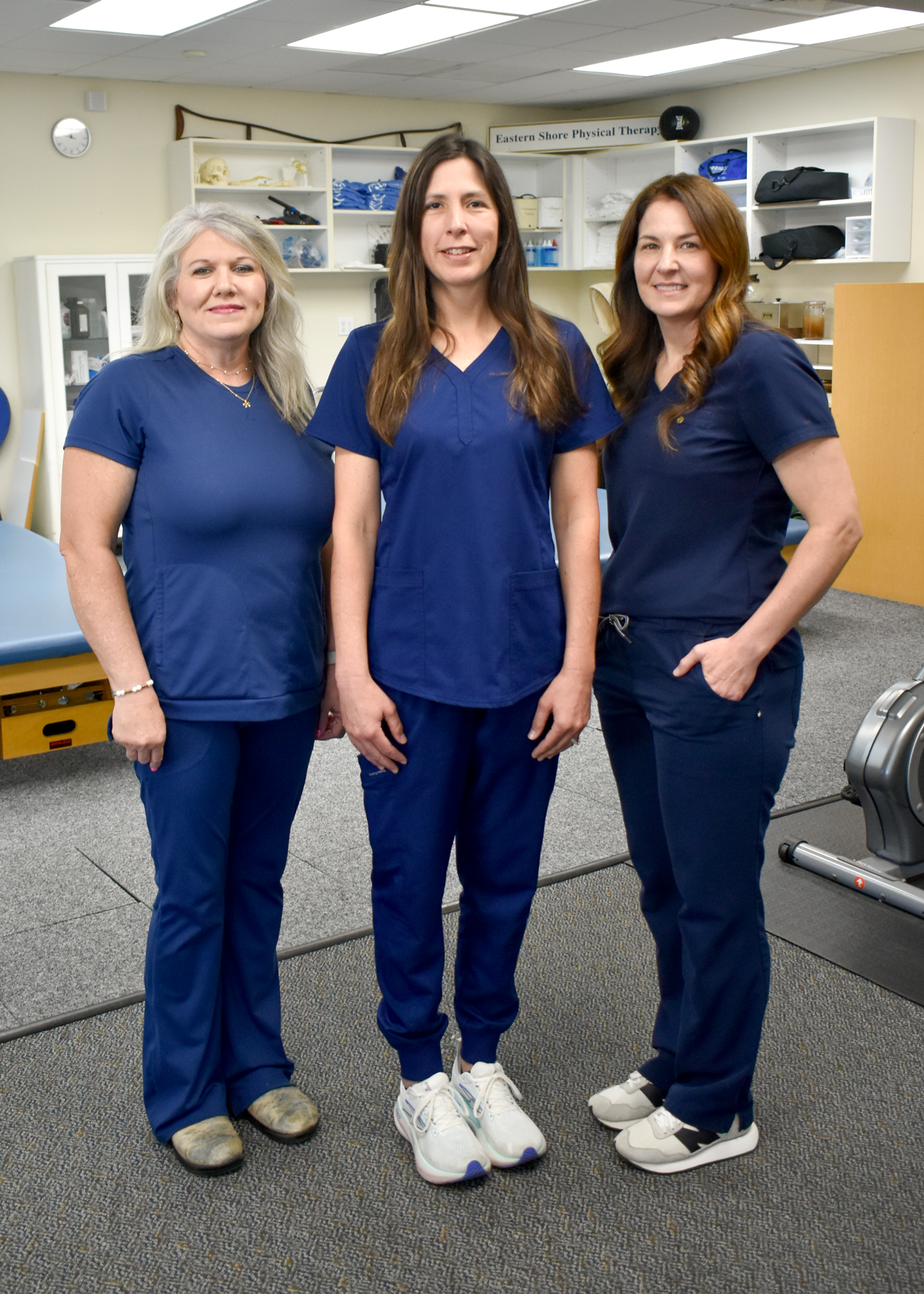 Three women wearing blue medical scrubs standing in a medical or therapy room with shelves and equipment in the background.