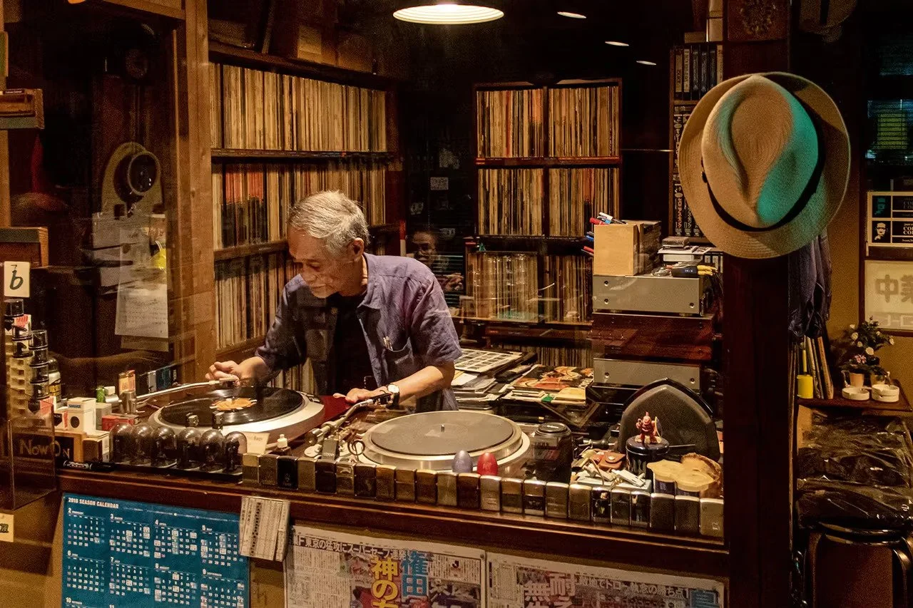A person DJing in a room filled with vinyl records, with shelves of records, a turntable, a mirror, and various items on the counter.