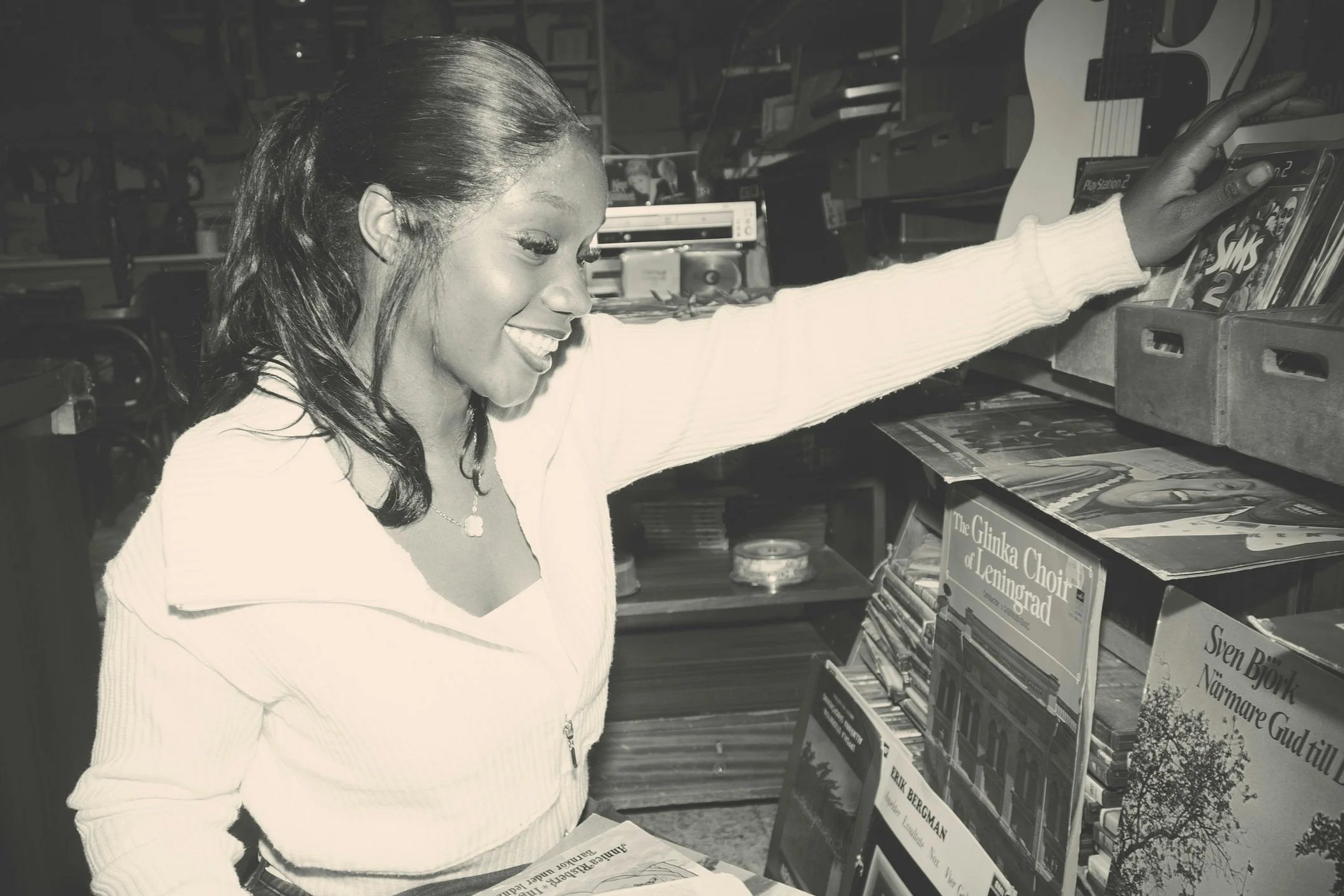 A woman with dark, wavy hair in a ponytail, wearing a light-colored sweater and a necklace, is smiling as she reaches for books on a shelf in a bookstore or library.