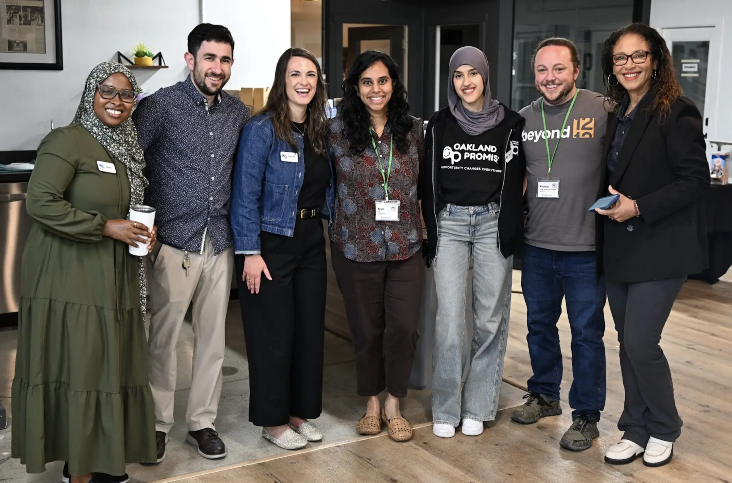 Group of eight diverse people standing indoors, smiling for a photo, wearing casual and business attire, some with conference badges.