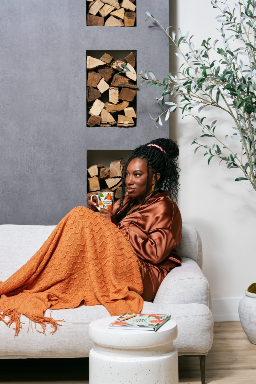Woman sitting on a sofa holding a coffee mug, wrapped in an orange blanket, with stacked firewood in the background and a large plant nearby.