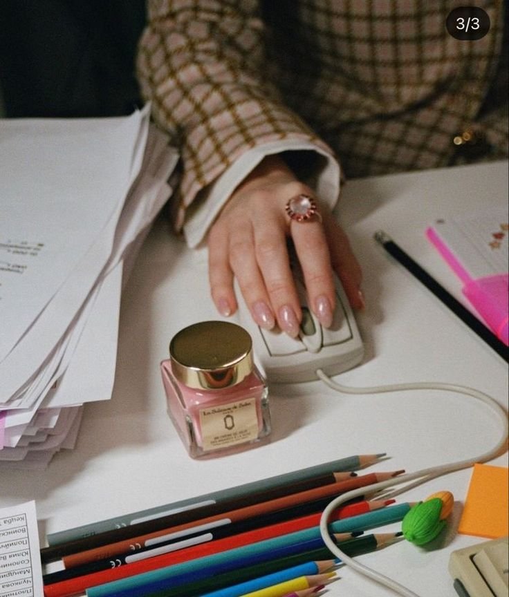 A person's hand with manicured nails and a large ring on the index finger, resting on a smartphone on a cluttered desk. The desk has papers, colorful pens, a nail polish bottle, a notebook, and a green earphone.