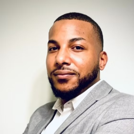 Portrait of a man with short hair and a beard, wearing a gray suit jacket and white shirt, smiling slightly against a plain background.