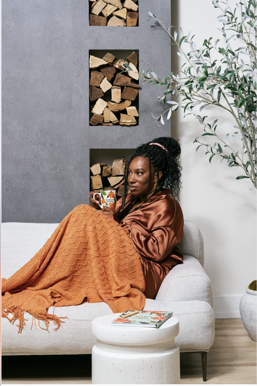 A woman with curly hair and a pink headband sits on a gray couch, wrapped in an orange blanket, holding a colorful mug. There are wooden logs in rectangular cutouts on a gray textured wall behind her, a leafy plant to her right, and a white coffee table with a magazine in front of her.