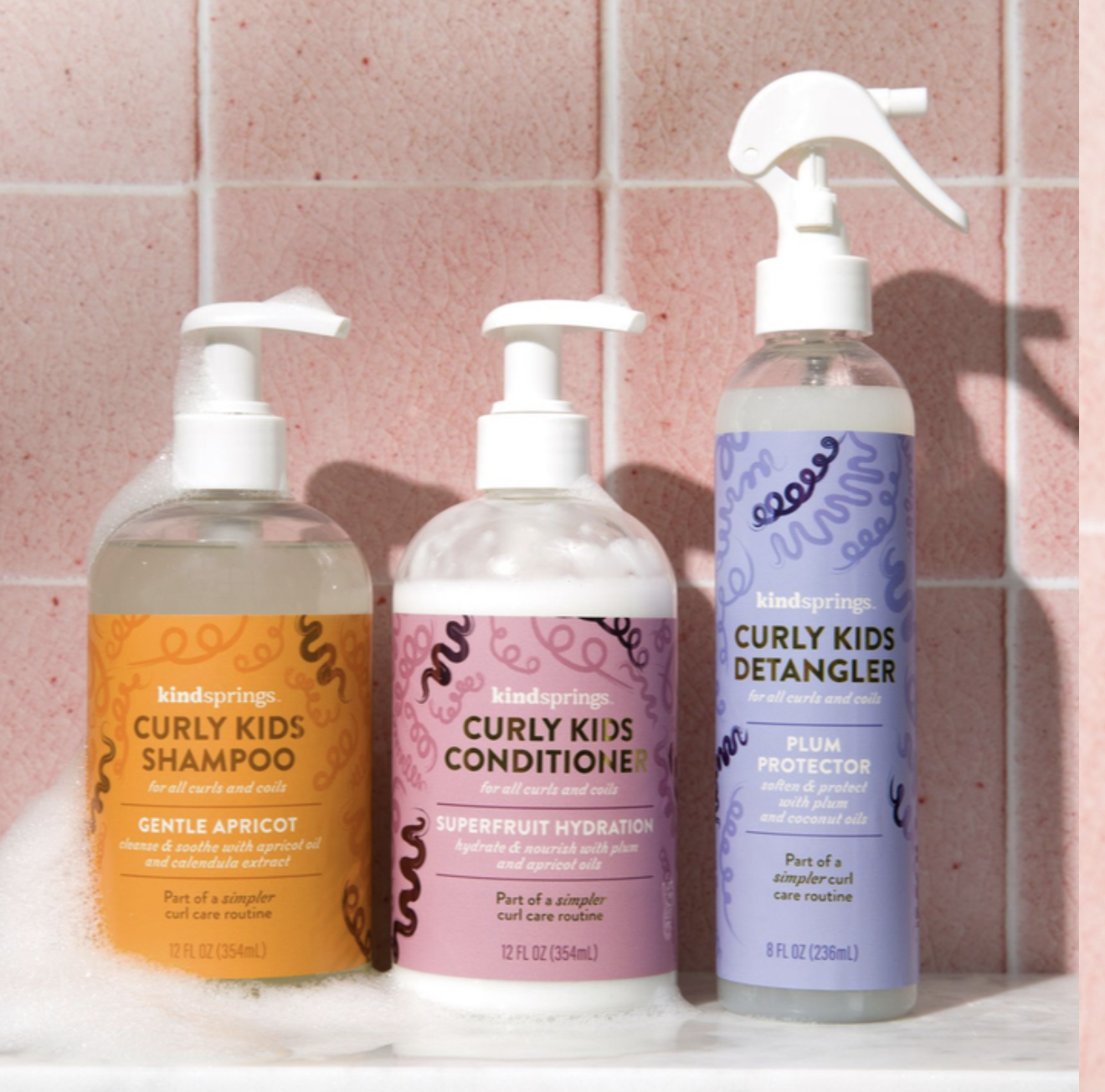 Three bottles of Kindsprings curly kids hair care products, including shampoo, conditioner, and detangler, placed on a white surface in front of pink tiled wall.