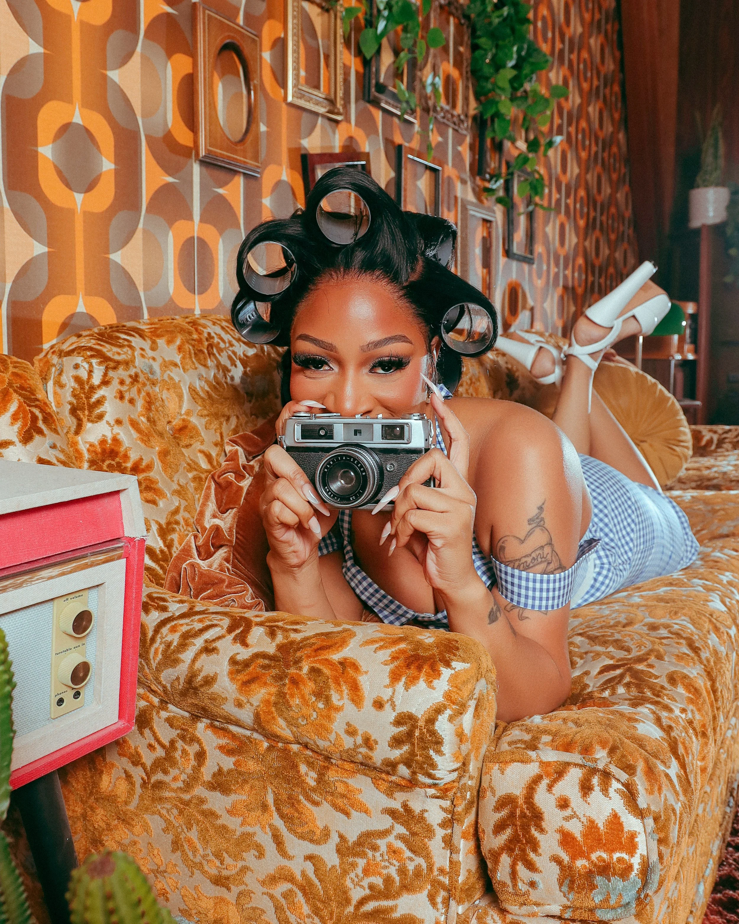 Woman with hair rollers, smiling, holding a vintage camera on a patterned orange and brown couch in a retro-style room.