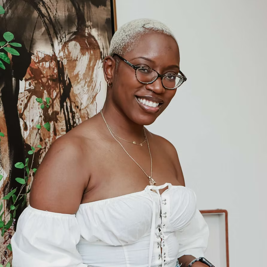 Smiling woman with short blonde hair, glasses, wearing a white off-the-shoulder top, standing indoors next to a painting and some greenery.