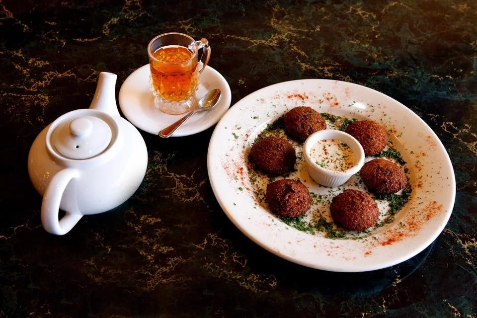 A white teapot, a small glass cup of tea with a spoon, and a white plate with six breaded meatballs around a small cup of dipping sauce, garnished with herbs and paprika on a dark marble table.