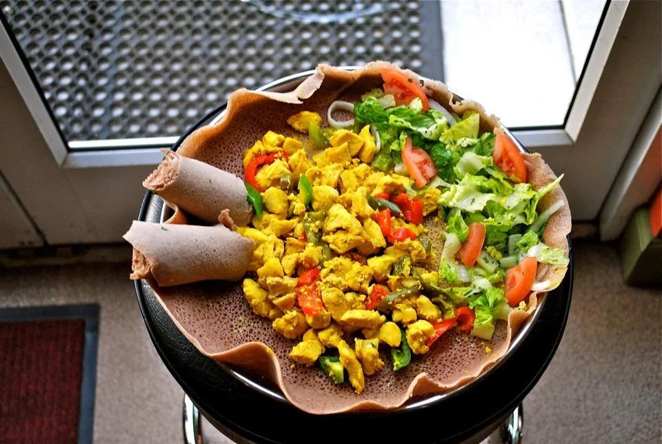 A plate of tofu scramble with chopped onions, tomatoes, lettuce, and two rolled crepes, on a black table near a glass door.