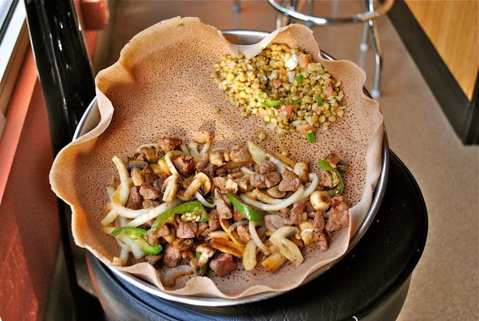 A bowl with two sections of cooked and seasoned chopped beef, onions, and green peppers, with a crispy edible bowl lining the rim, topped with two different types of cooked grains or lentils.
