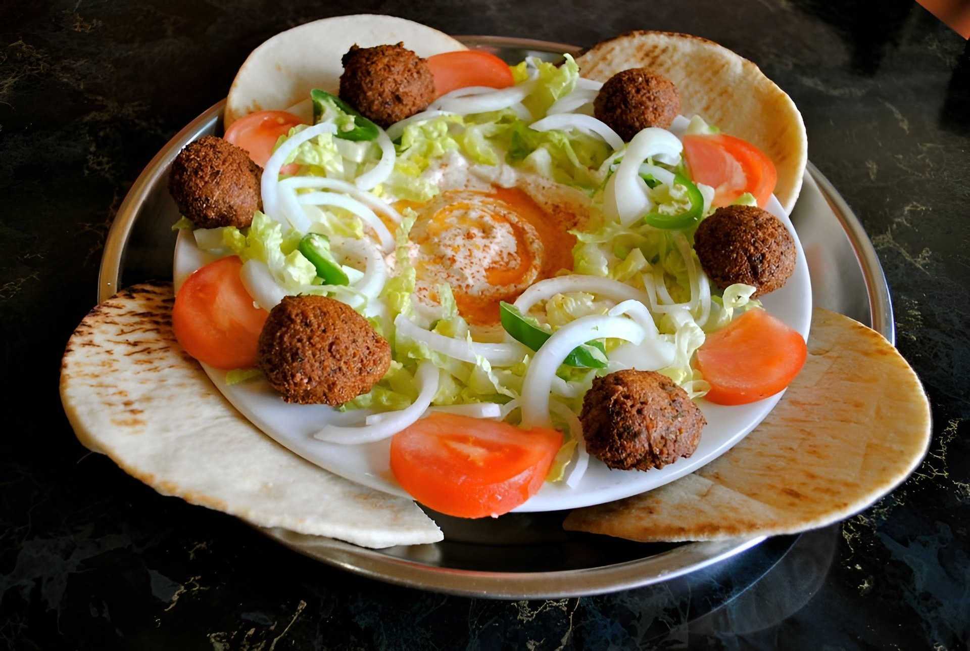 A plate of food with fried falafel balls, sliced tomatoes, onion rings, lettuce, a boiled egg, and pita chips, arranged around a bed of lettuce with a soft boiled egg in the center, served on a silver tray with a black marble background.