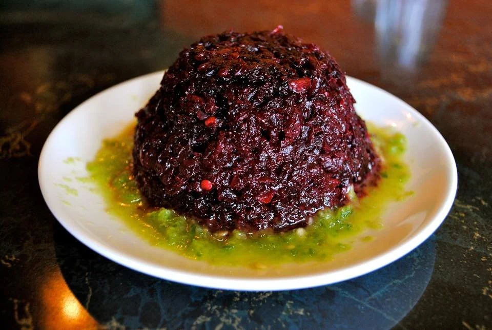 A white plate holding a dark, saucy meat dish with green sauce underneath, on a dark surface.