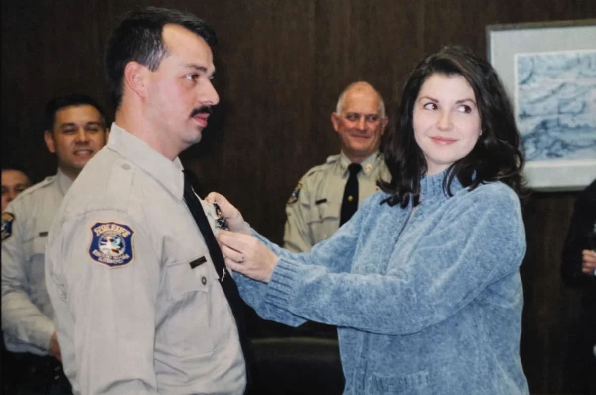 A woman awards a badge to a male police officer in a uniform while smiling, with other officers watching in the background.
