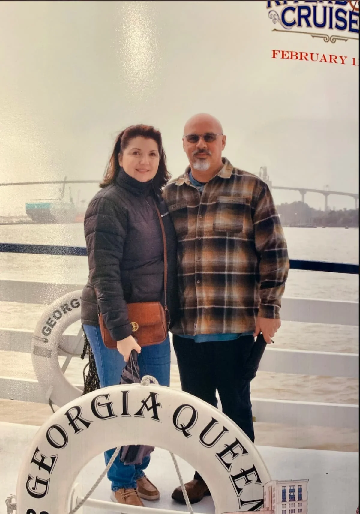 A man and a woman standing together on a cruise ship deck with a city skyline and bridge in the background. The woman wears a black jacket and has a brown purse, while the man wears a plaid shirt and glasses. They are smiling and posing for the photo.
