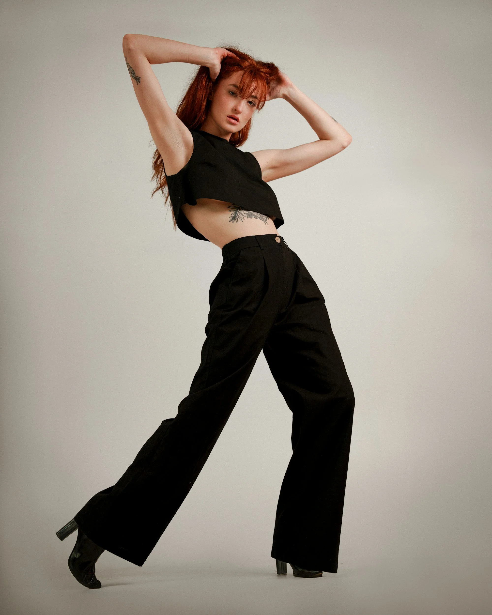 A woman with red hair and tattoos is posing against a plain background, wearing a black cropped top, high-waisted black pants, and black high-heeled boots.