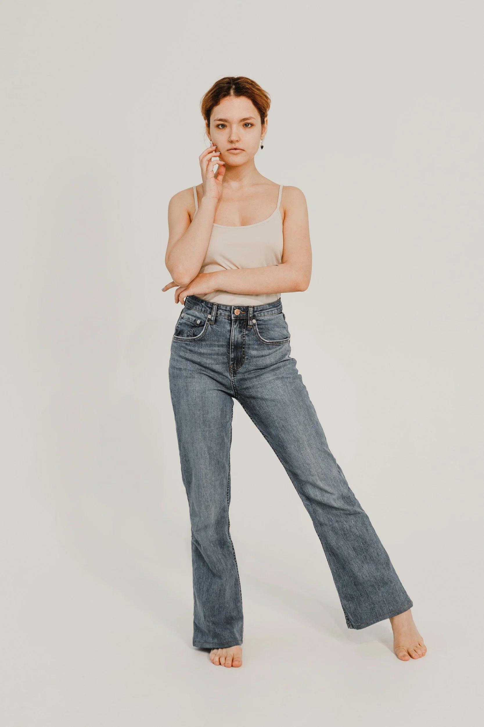 A young woman with short brown hair standing against a plain white background, wearing a beige spaghetti strap top and high-waisted blue jeans, with one arm crossed and the other touching her face.