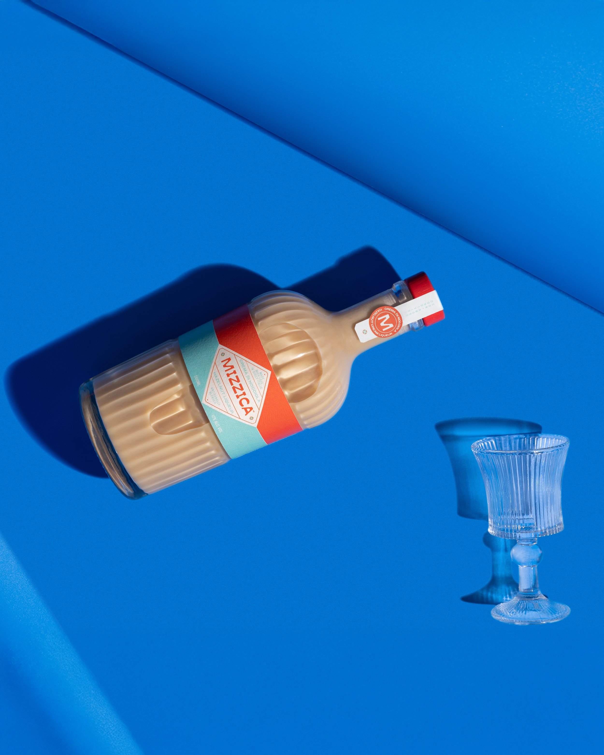 A bottle of beige-colored beverage with a red cap lying on a blue surface with a blue wall in the background, next to a clear glass goblet. Photography by Lila Marvell www.lilamarvell.com.au