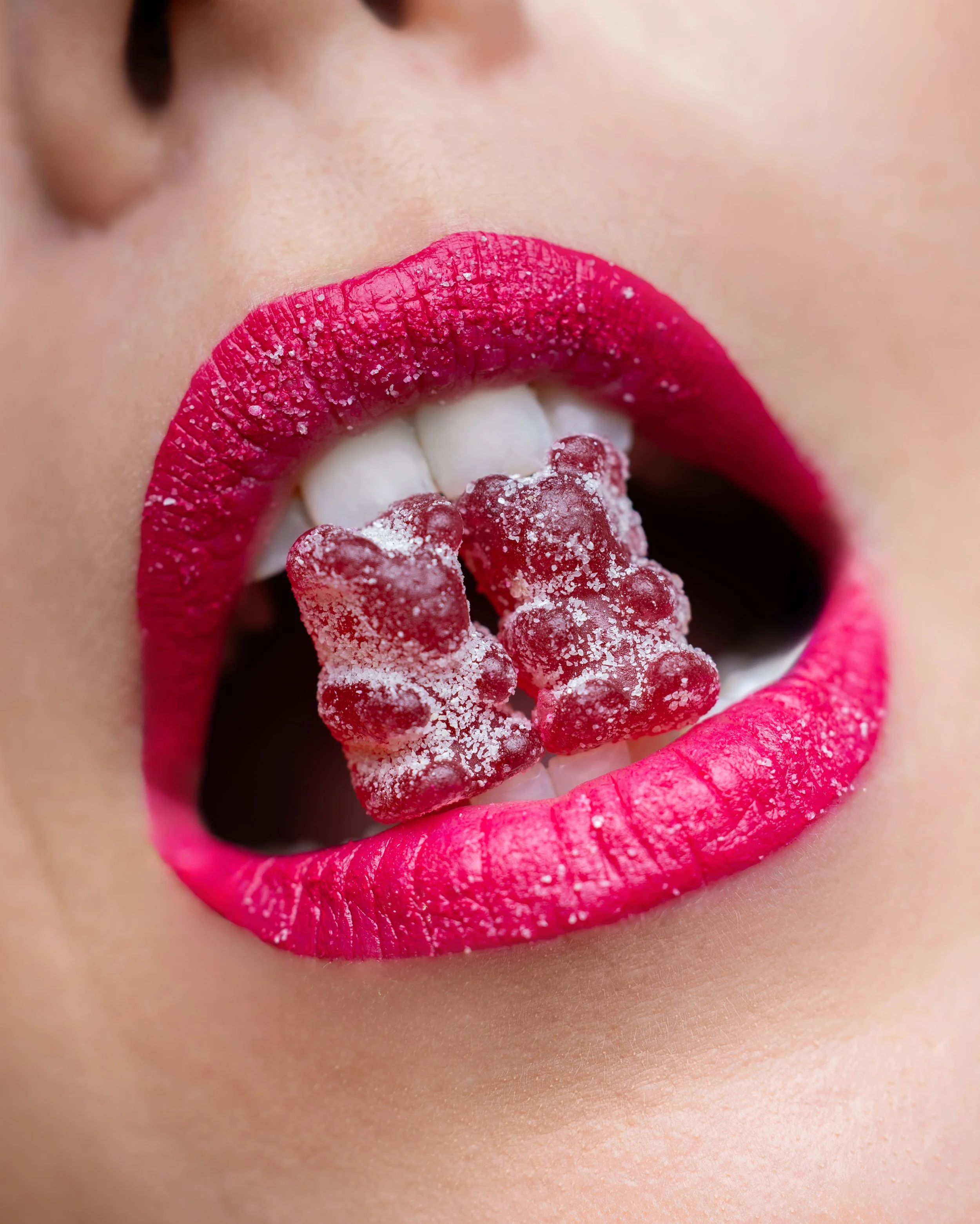 Close-up of a person's lips with bright pink lipstick, holding red gummy bear candies with sugar on them in mouth. Photography by Lila Marvell www.lilamarvell.com.au