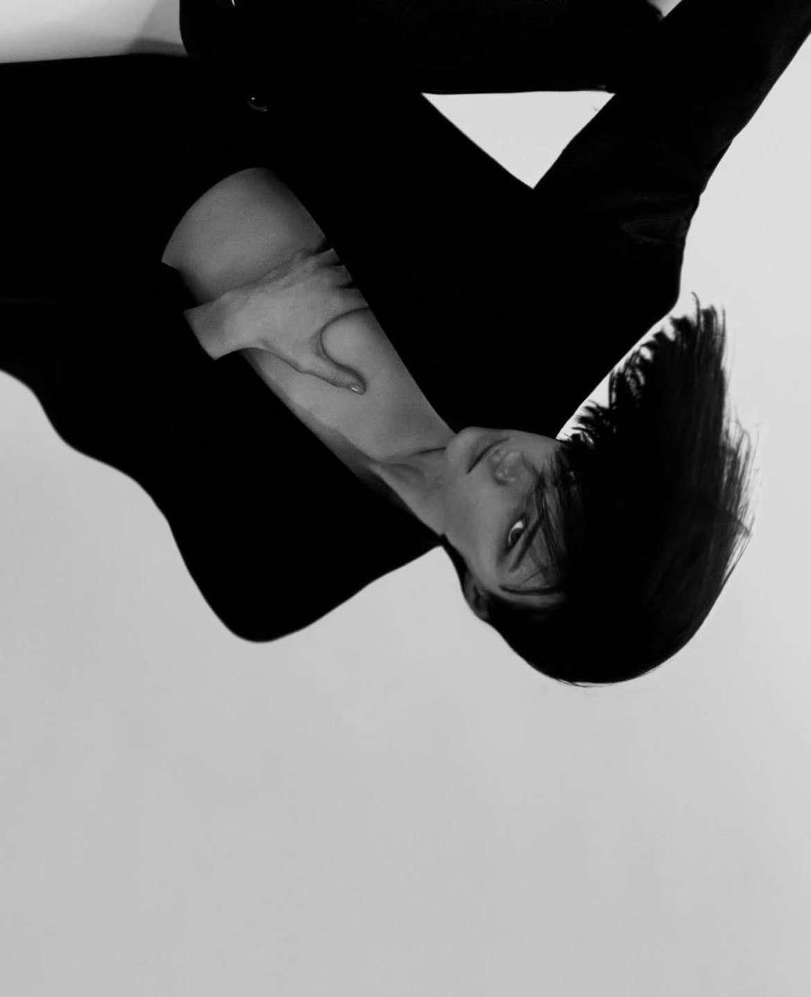 Black and white photo of a woman with short hair, making a playful face with her tongue out, while holding her leg with both hands.