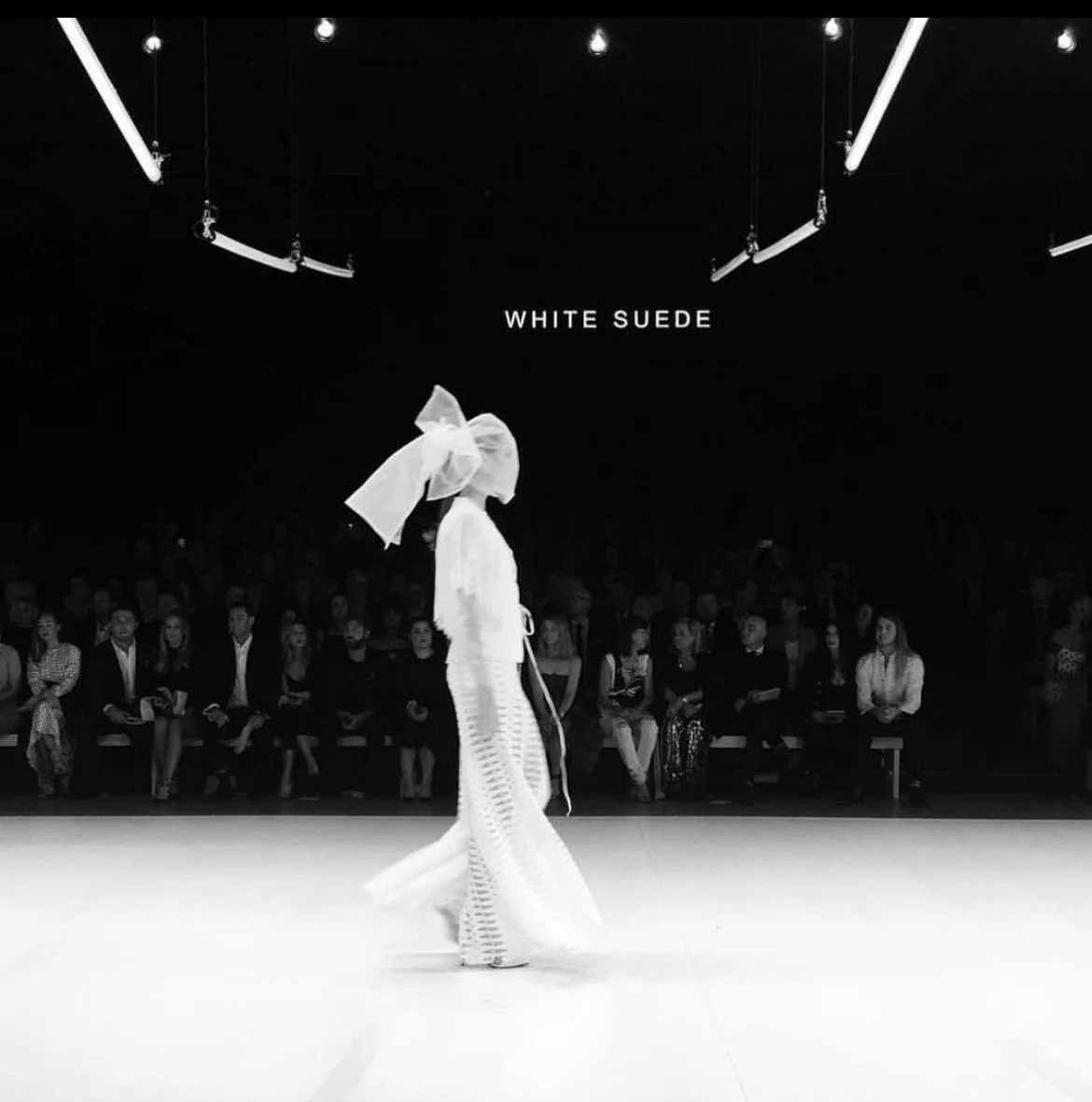 A fashion runway show featuring a model in white clothing with a large bow on her head, walking on a white floor with an audience seated in the background and overhead lighting fixtures.