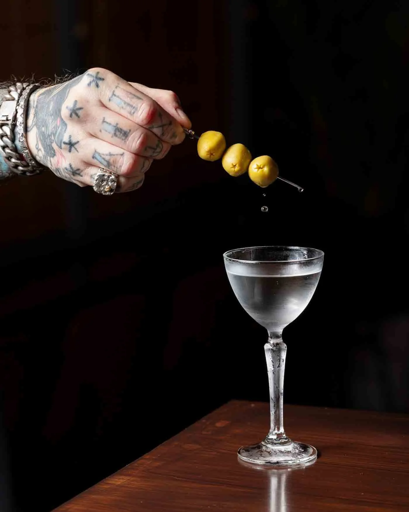 A hand with tattoos holding a skewer of three yellow cherries above a glass of clear liquid on a wooden surface. Photography by Lila Marvell www.lilamarvell.com.au
