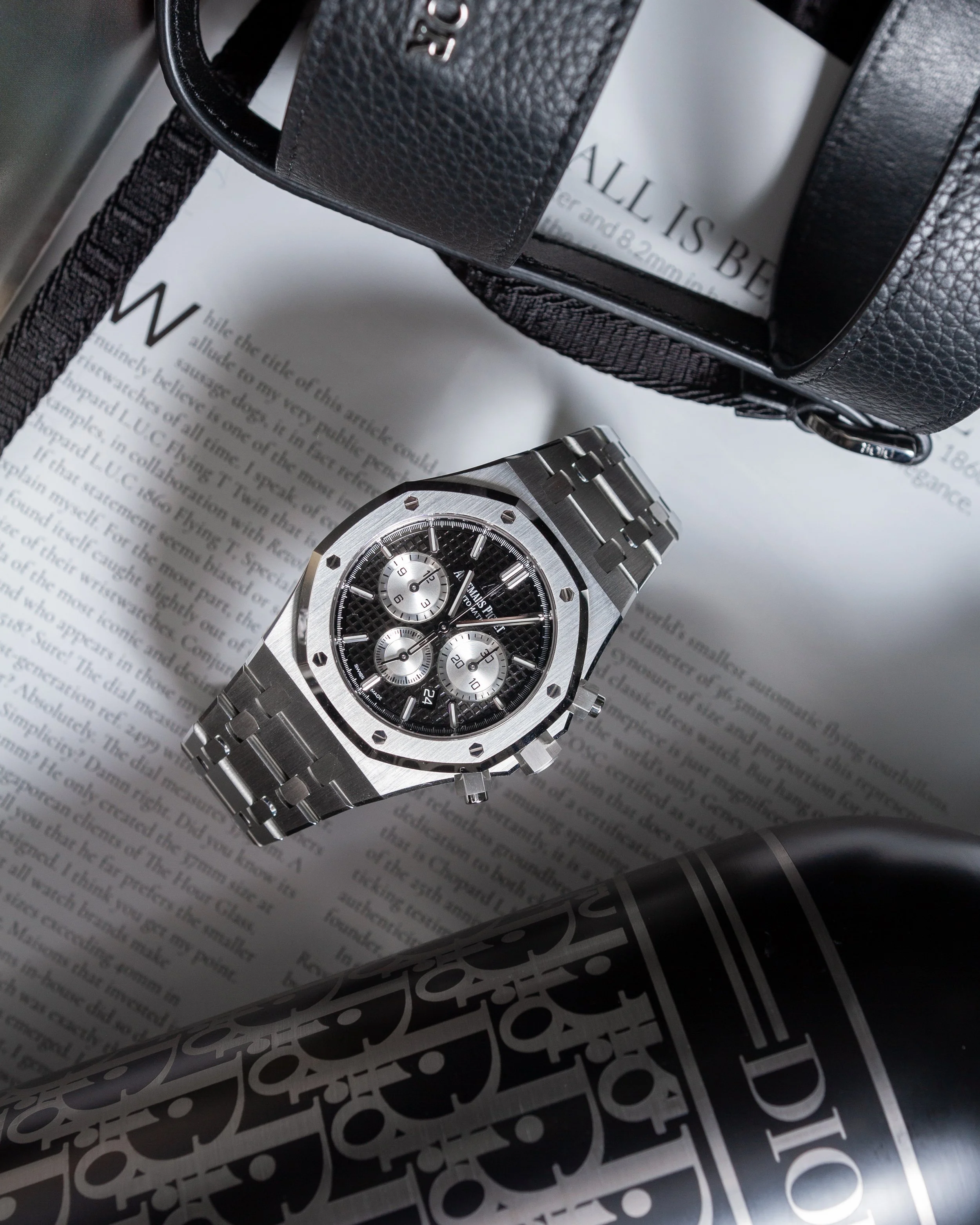 A silver wristwatch with a black face and subdials, placed on an open magazine or newspaper, next to a black Dior bottle and a black handbag. Photography by Lila Marvell www.lilamarvell.com.au